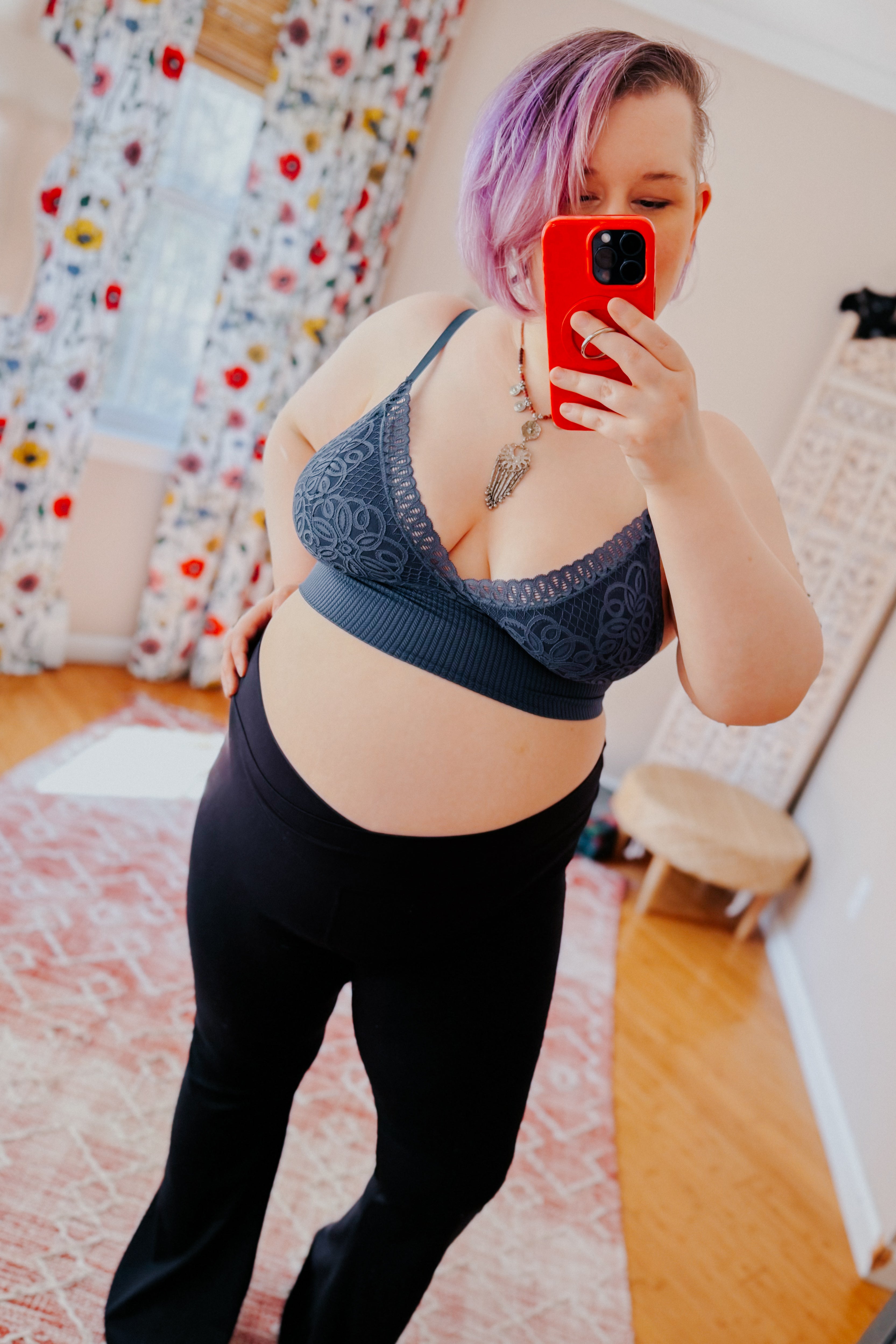 Woman with purple hair wearing dusty blue lace bralette and black pants taking selfie in sunlit room with floral curtains
