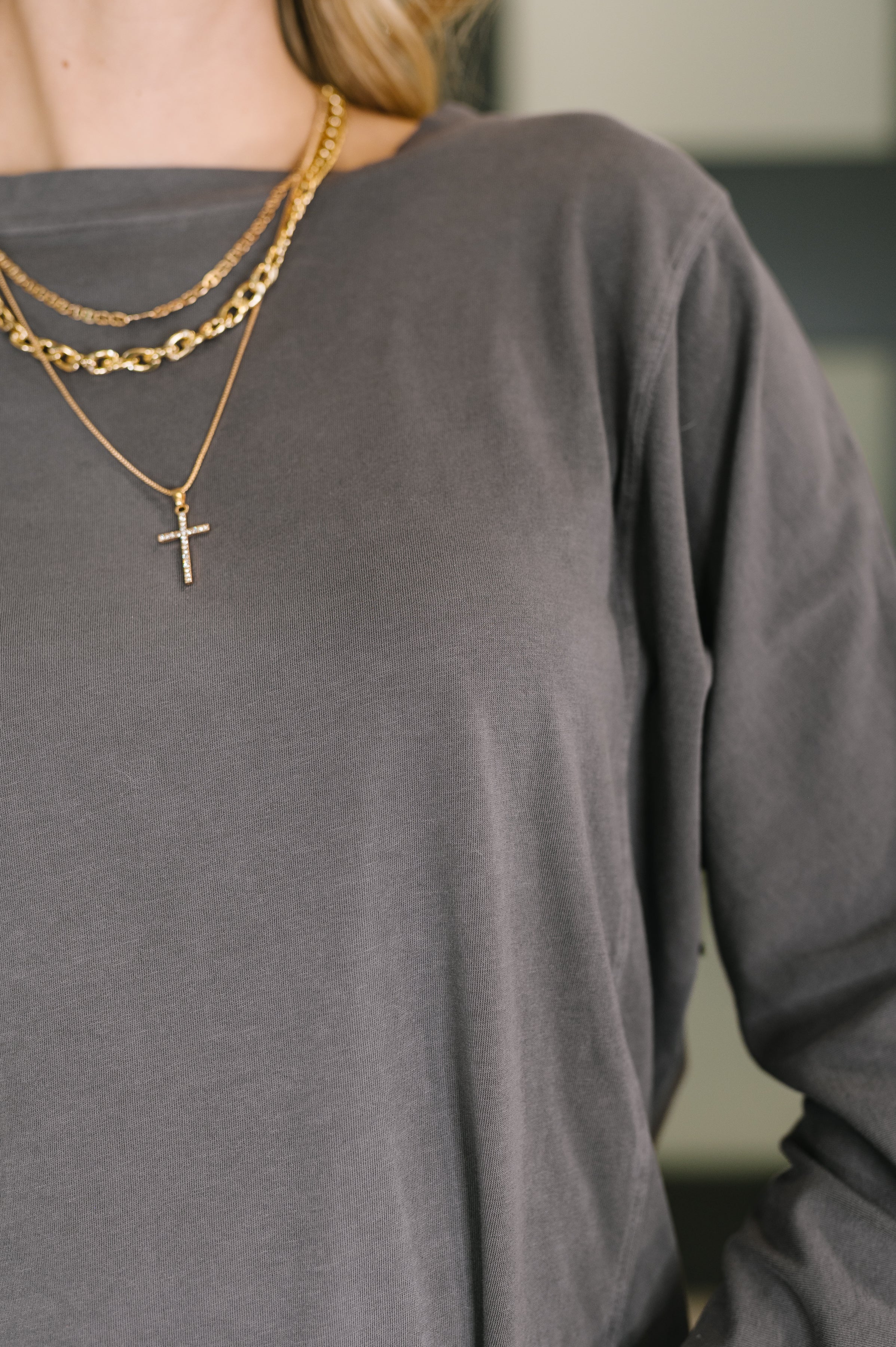 Close-up of woman wearing charcoal gray dolman sleeve top and layered gold chain necklaces with cross pendant indoors