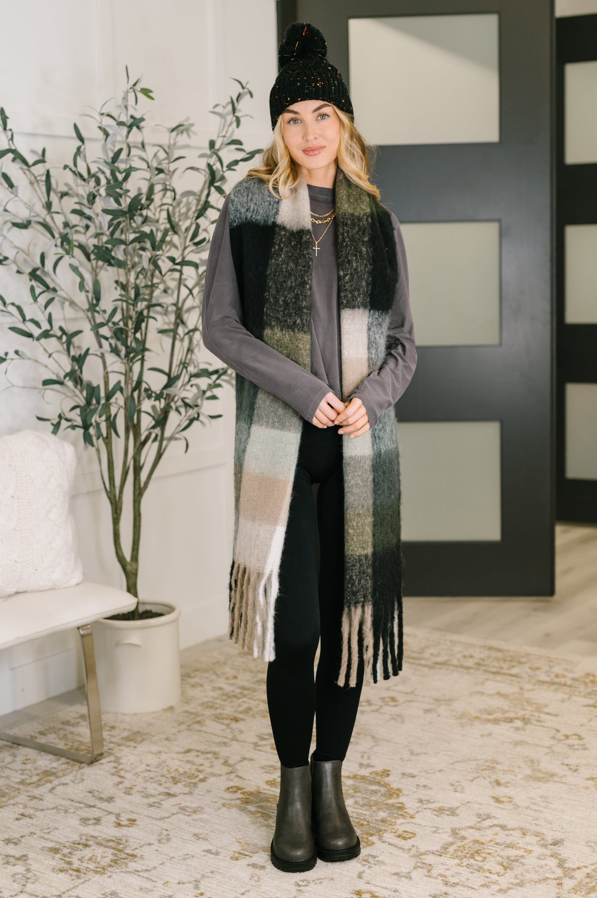 Woman wearing a grey dolman sleeve top with a fringed plaid scarf, black leggings, beanie, and ankle boots indoors