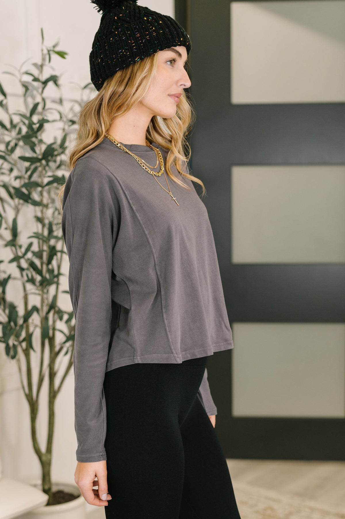 Woman wearing a grey dolman sleeve top with a relaxed draped fit, black beanie, layered necklaces and black pants indoors