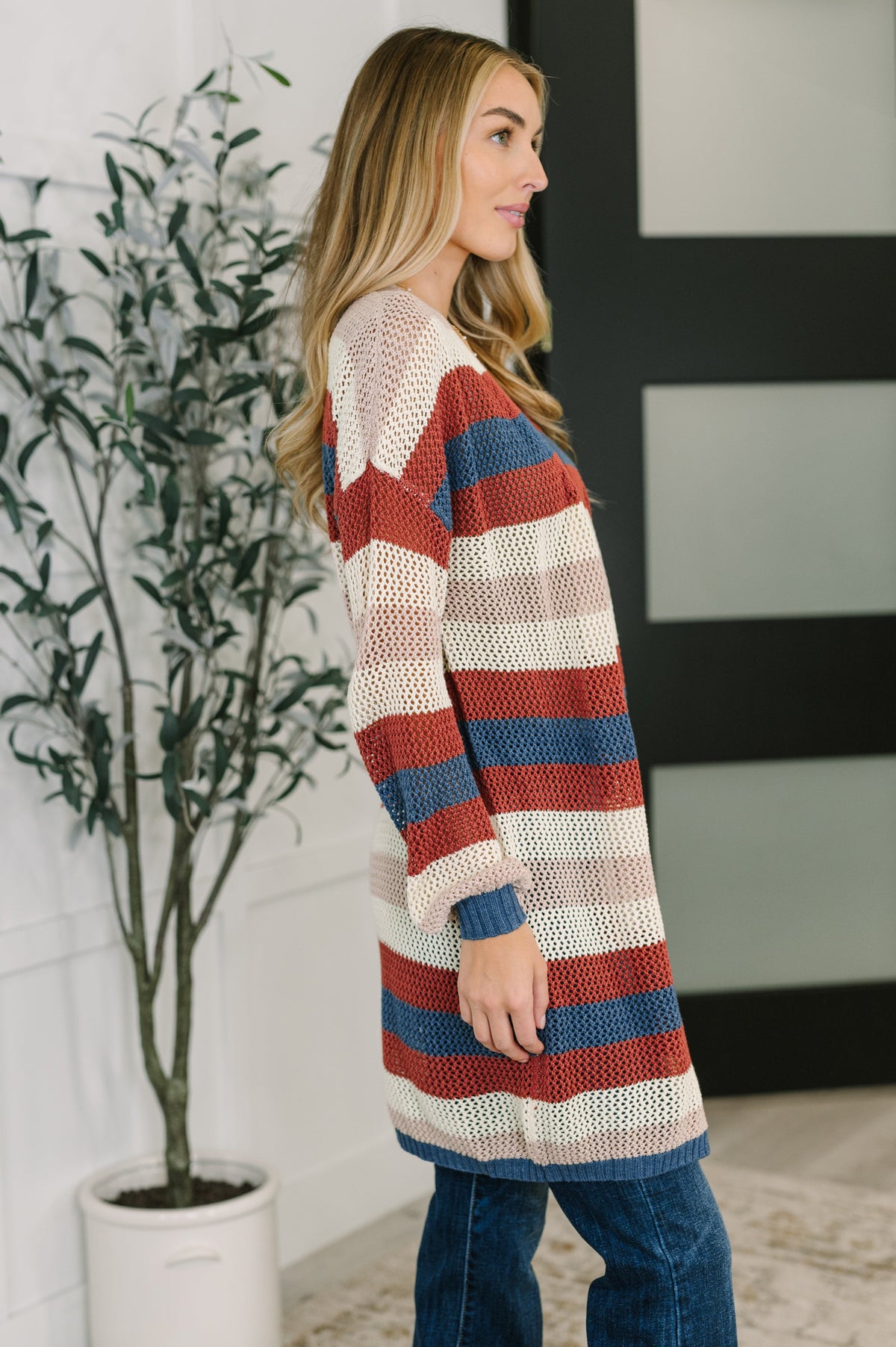 Woman in profile wearing a rust striped knit cardigan over jeans beside a potted plant in a modern home interior