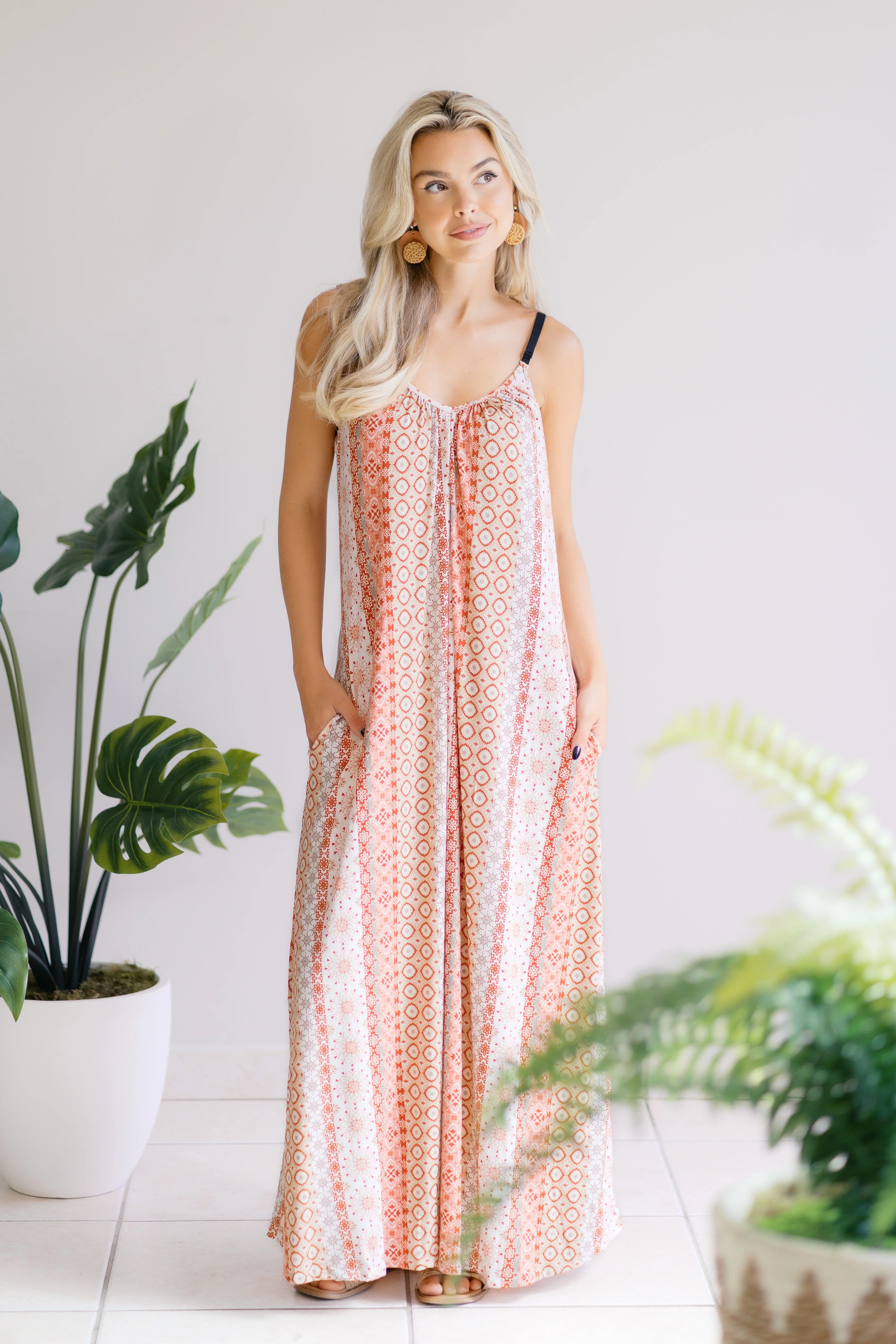 Blonde woman wearing patterned sleeveless maxi dress with pockets standing indoors near potted plants