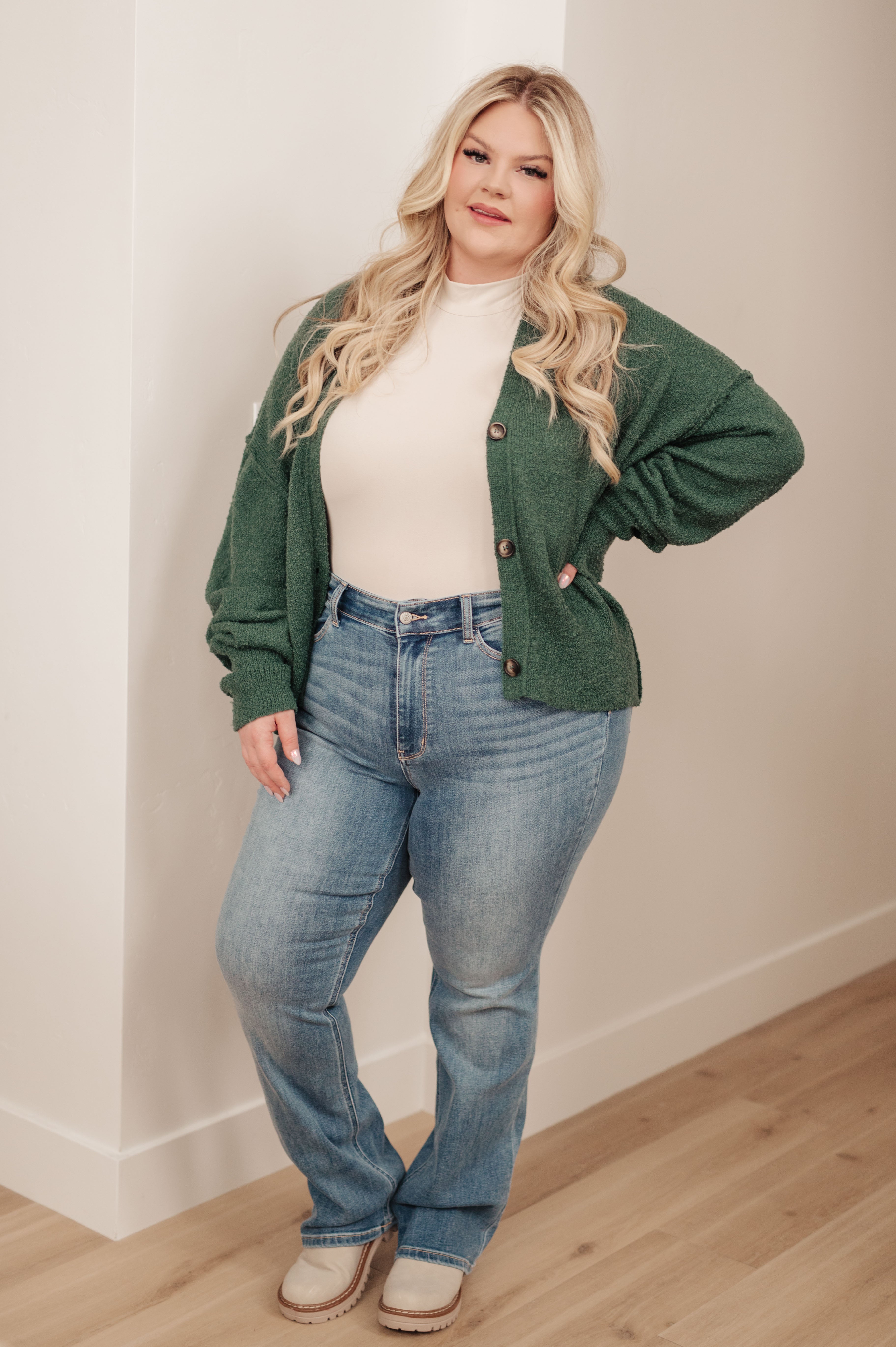 Plus size woman wearing a textured green button-up cardigan with light beige top and relaxed blue jeans indoors by white wall