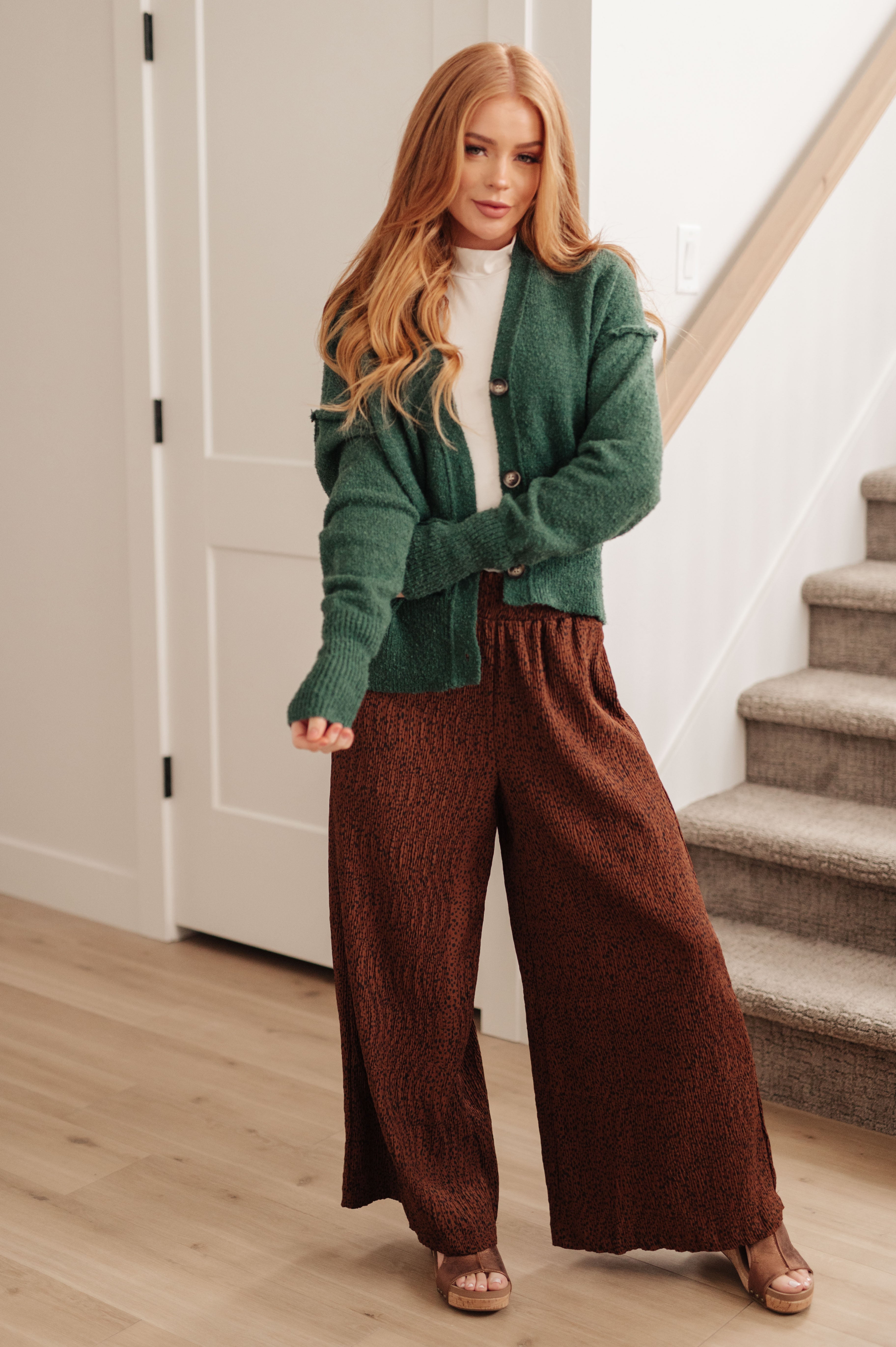 Woman with long blonde hair wearing forest green button-up cardigan and wide-leg brown pants indoors near staircase