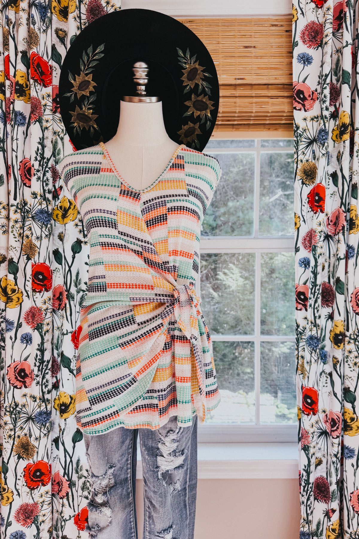 Mannequin wearing a blue striped faux wrap V neck sweater tank with distressed jeans and a black floral wide brim hat indoors