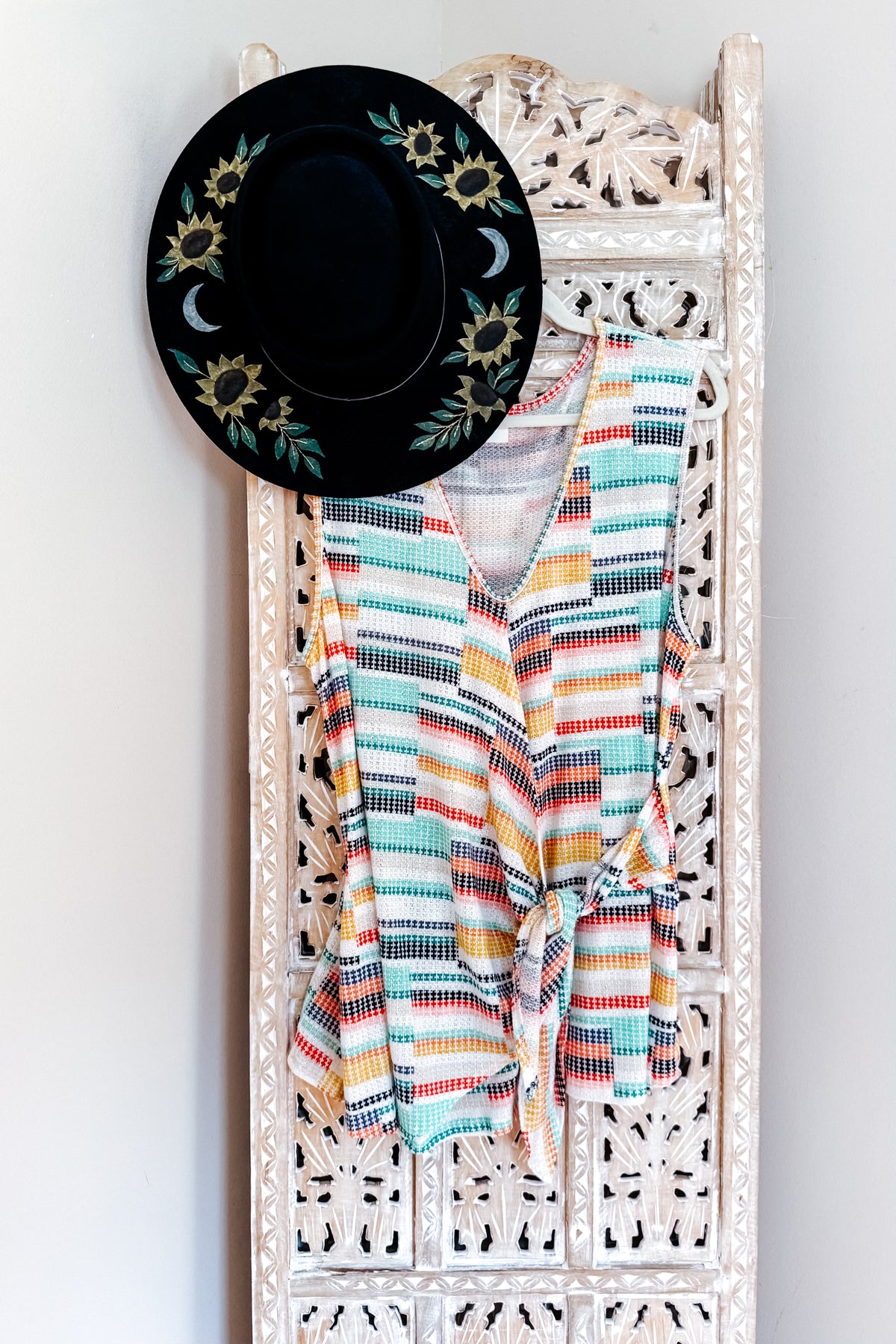Blue multicolored striped V neck faux wrap tank hanging on a white carved wall rack beneath a black floral embroidered hat