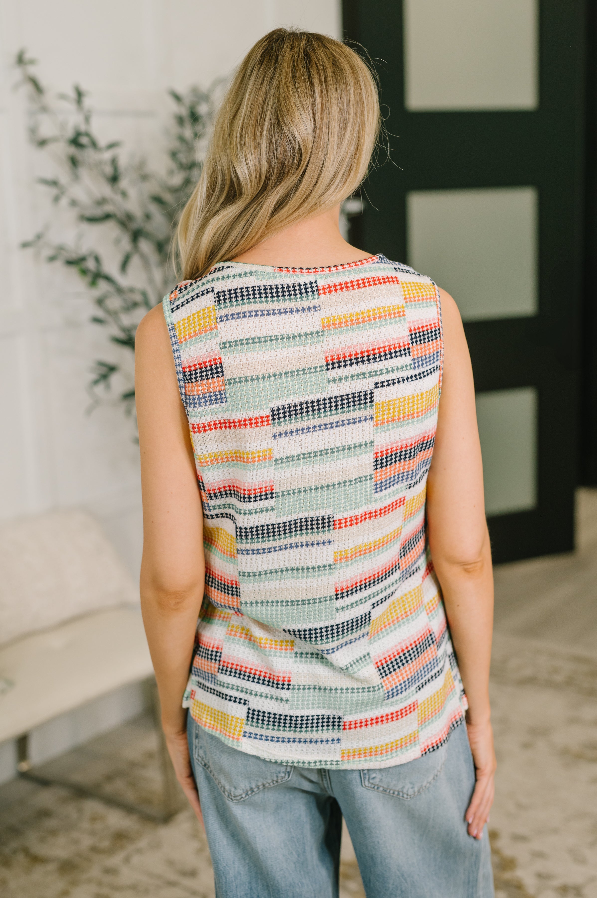 Back view of woman wearing sleeveless multicolor geometric print top paired with light wash jeans indoors
