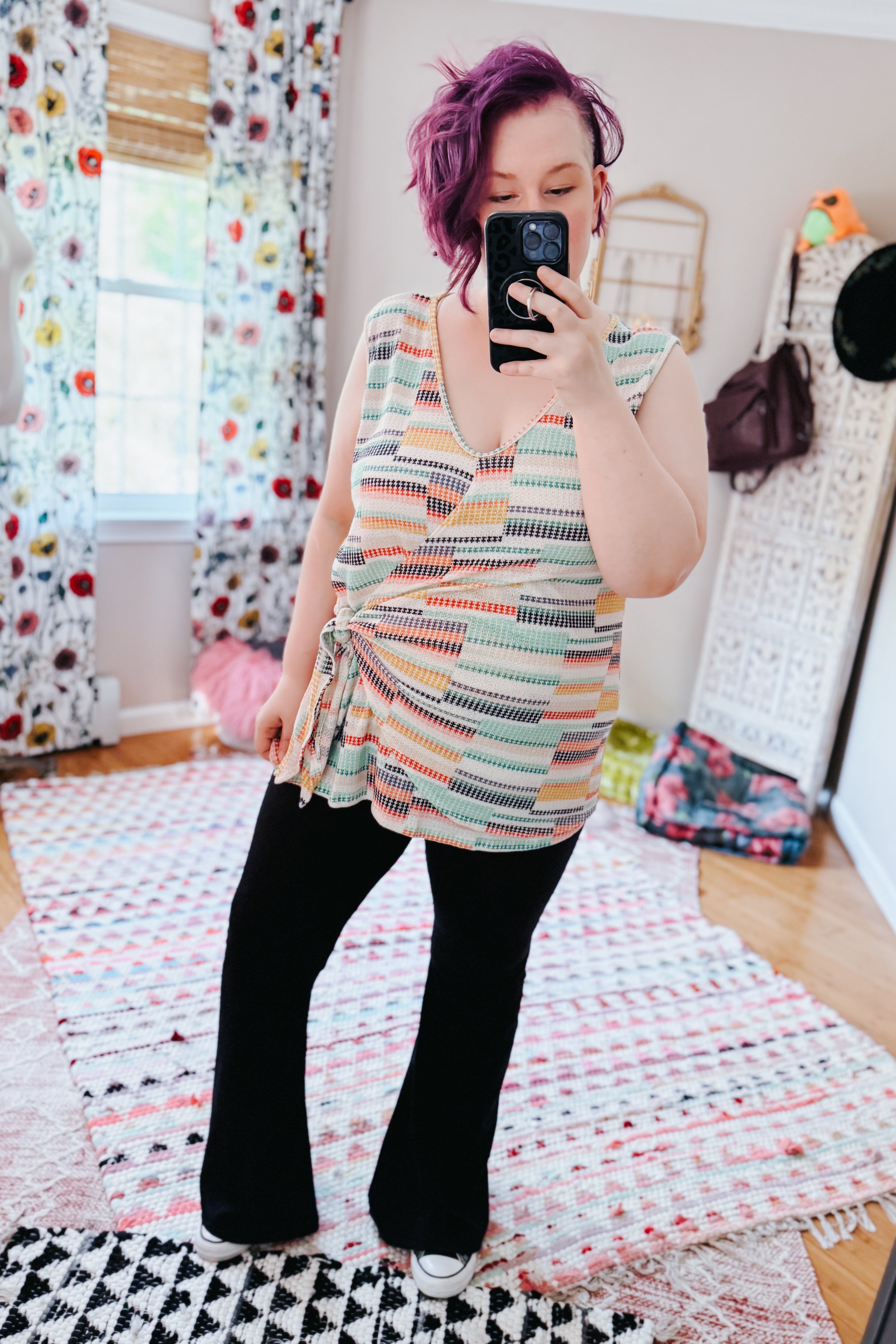 Woman with purple hair wearing a patterned sleeveless V-neck knit top and black pants in a bright eclectic room