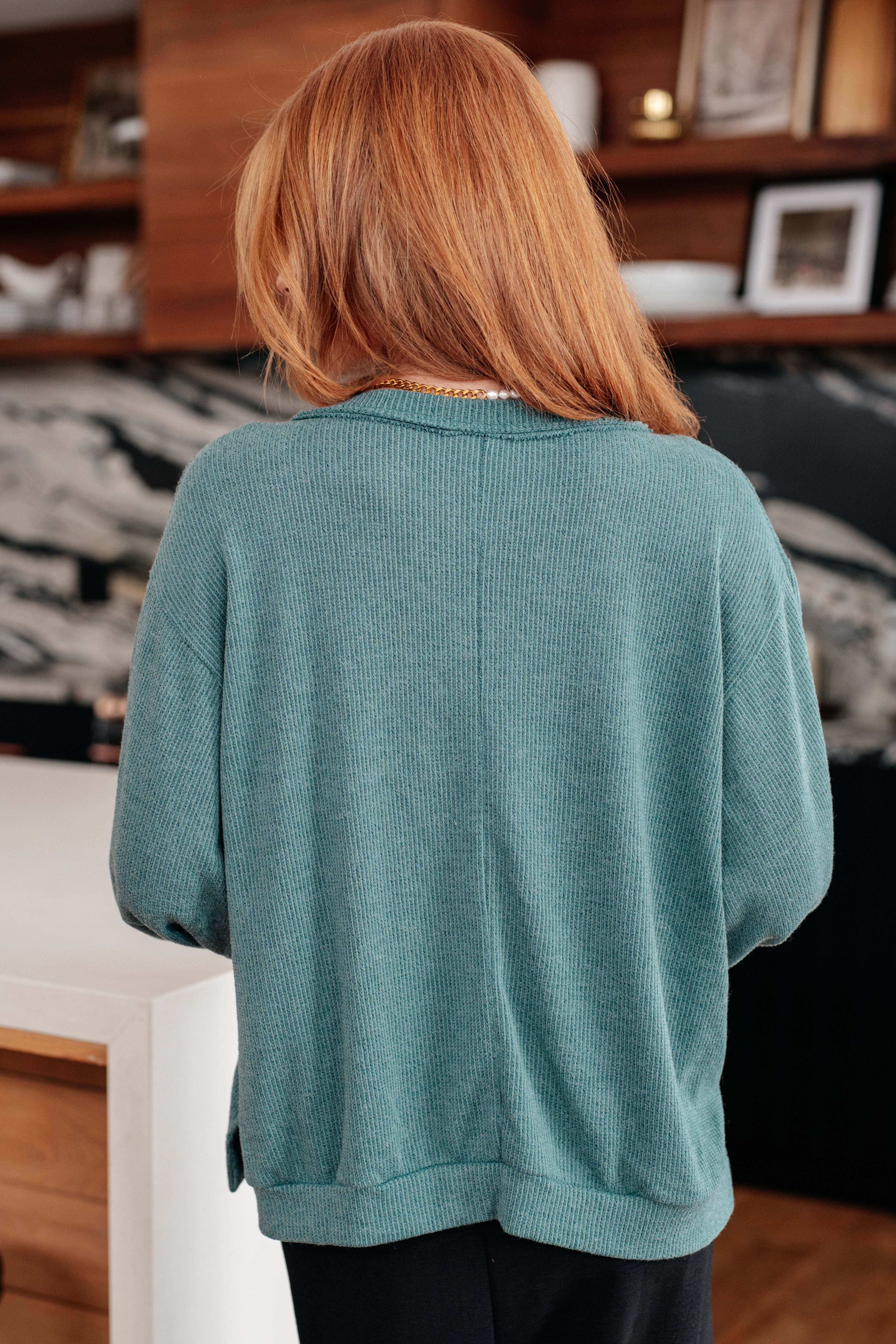 Woman with red hair facing away wearing a teal knit sweater in a modern kitchen setting