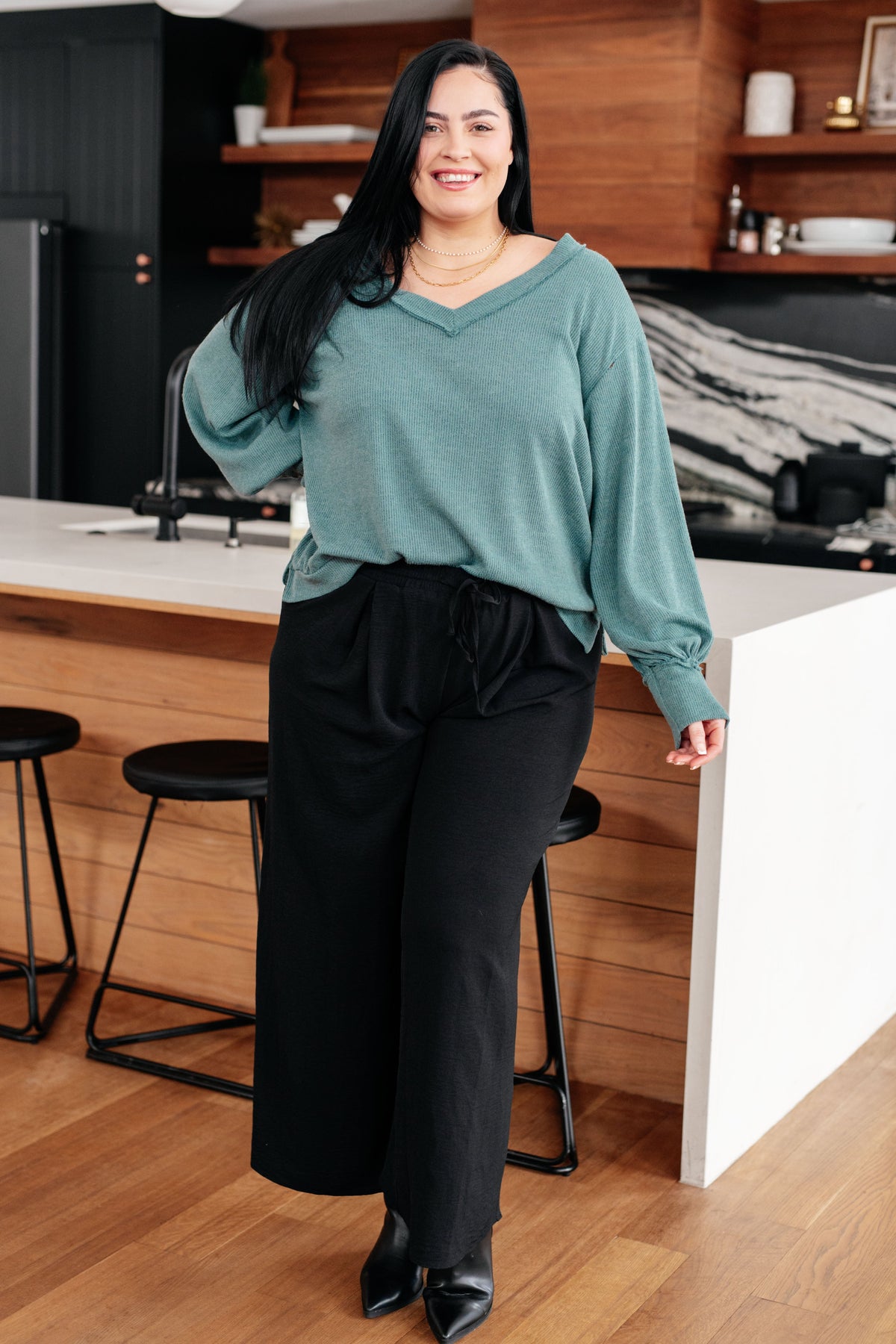 Woman wearing a blue brushed knit V-neck top with balloon sleeves and black wide-leg pants in a modern kitchen