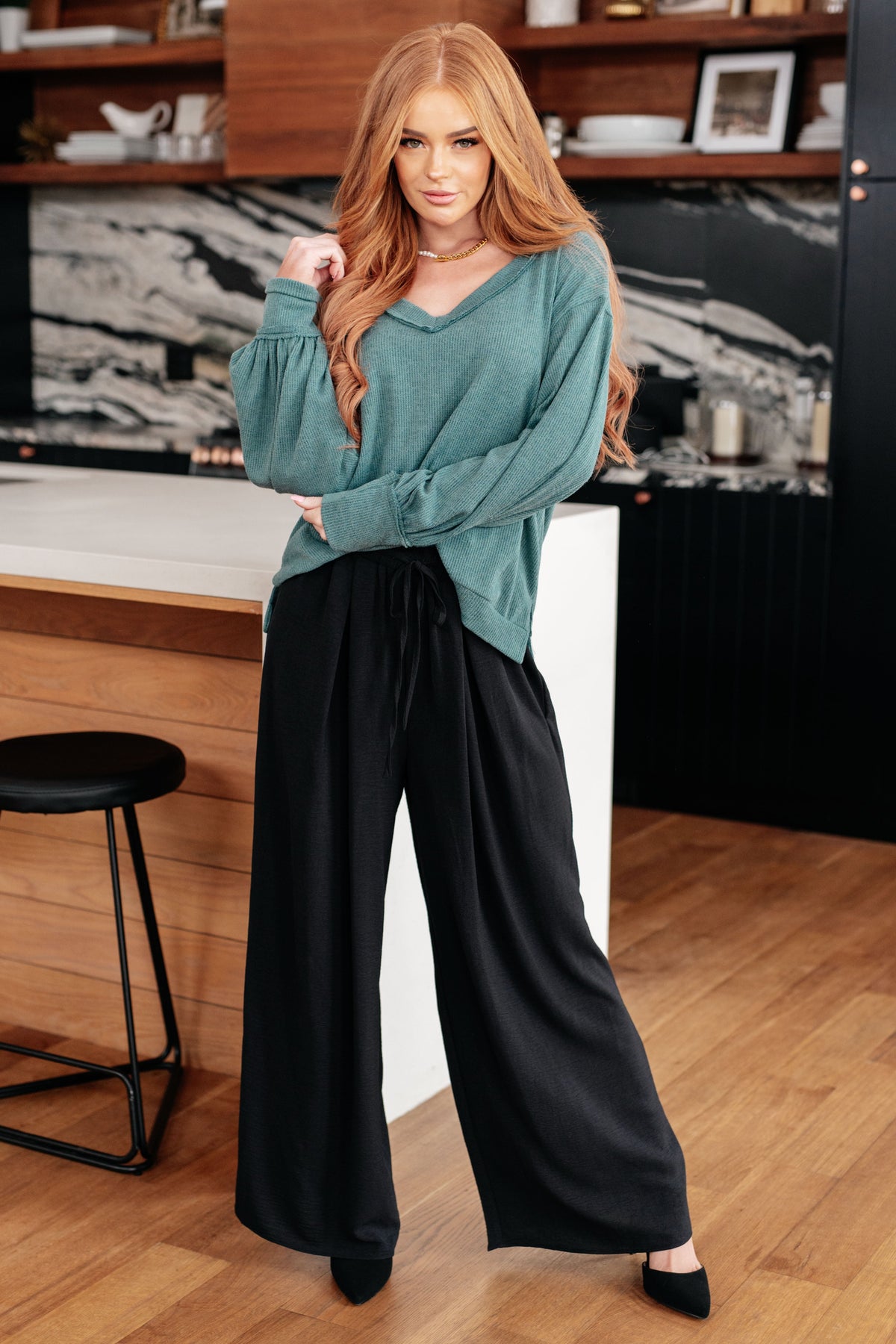 Woman wearing a blue brushed knit V-neck top with balloon sleeves and black wide-leg pants in a modern kitchen