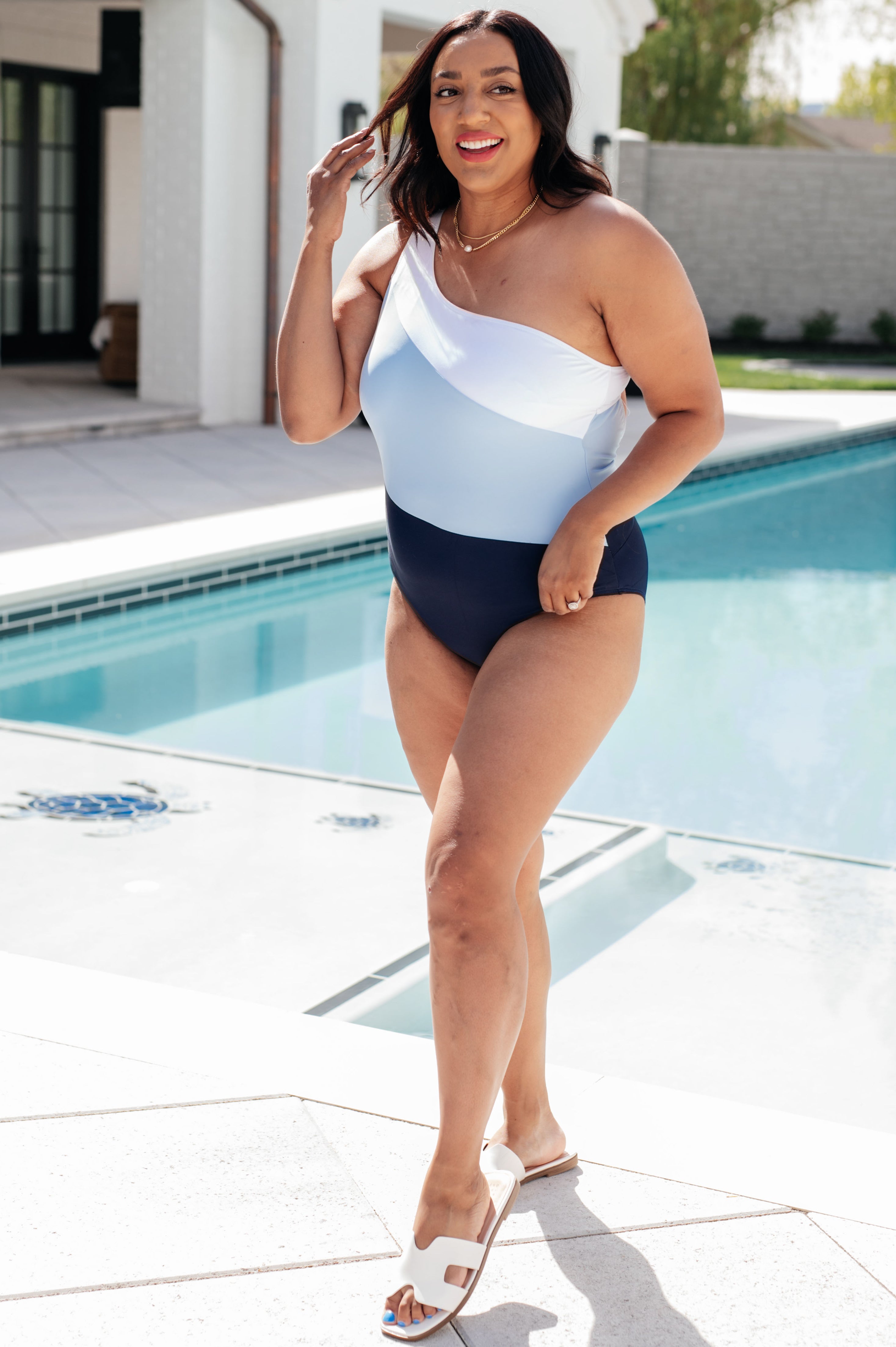 Curvy woman wearing a blue asymmetrical one-shoulder swimsuit standing beside an outdoor pool in white sandals