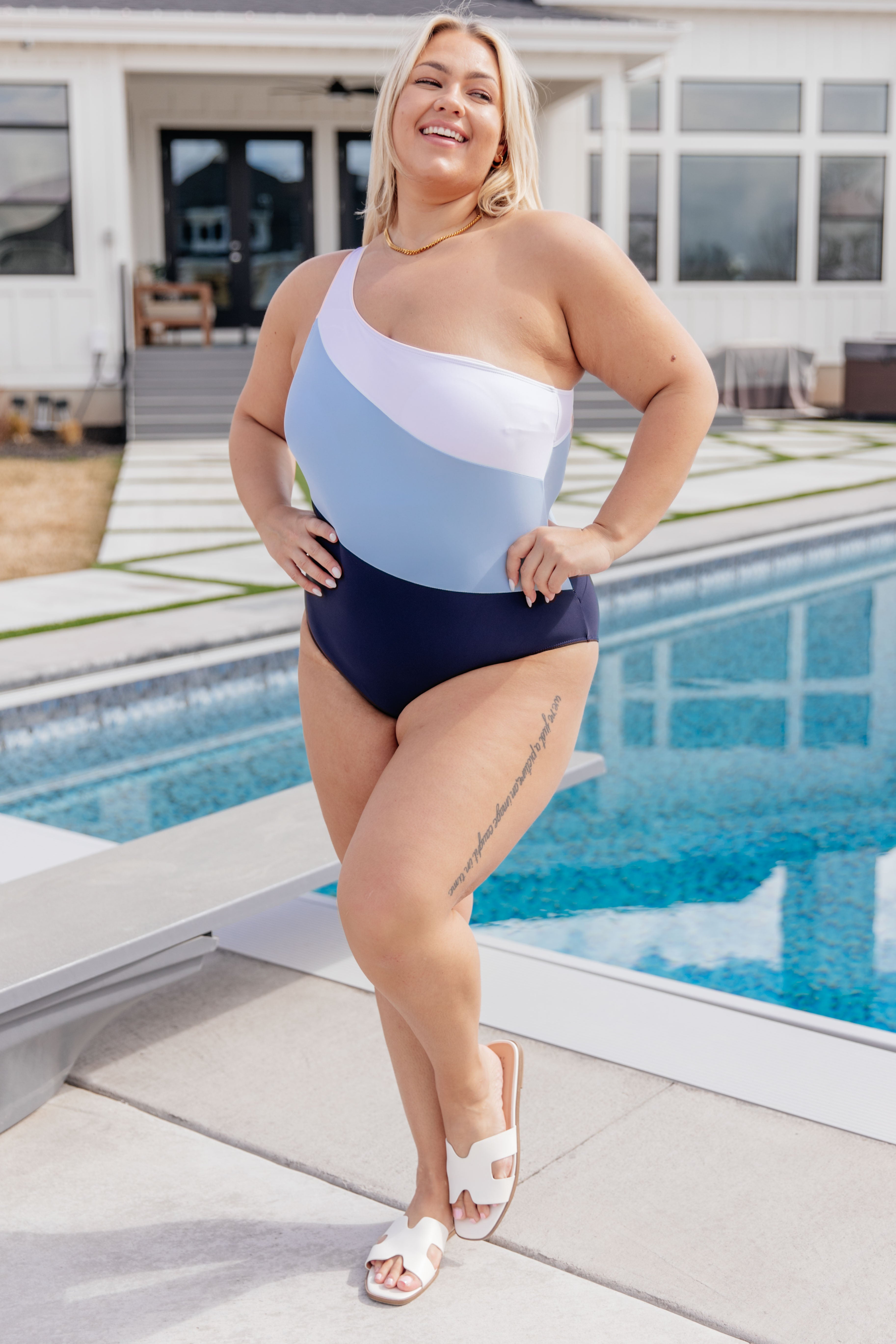 Smiling woman wearing a blue and white asymmetrical one-shoulder swimsuit standing near a pool in white sandals