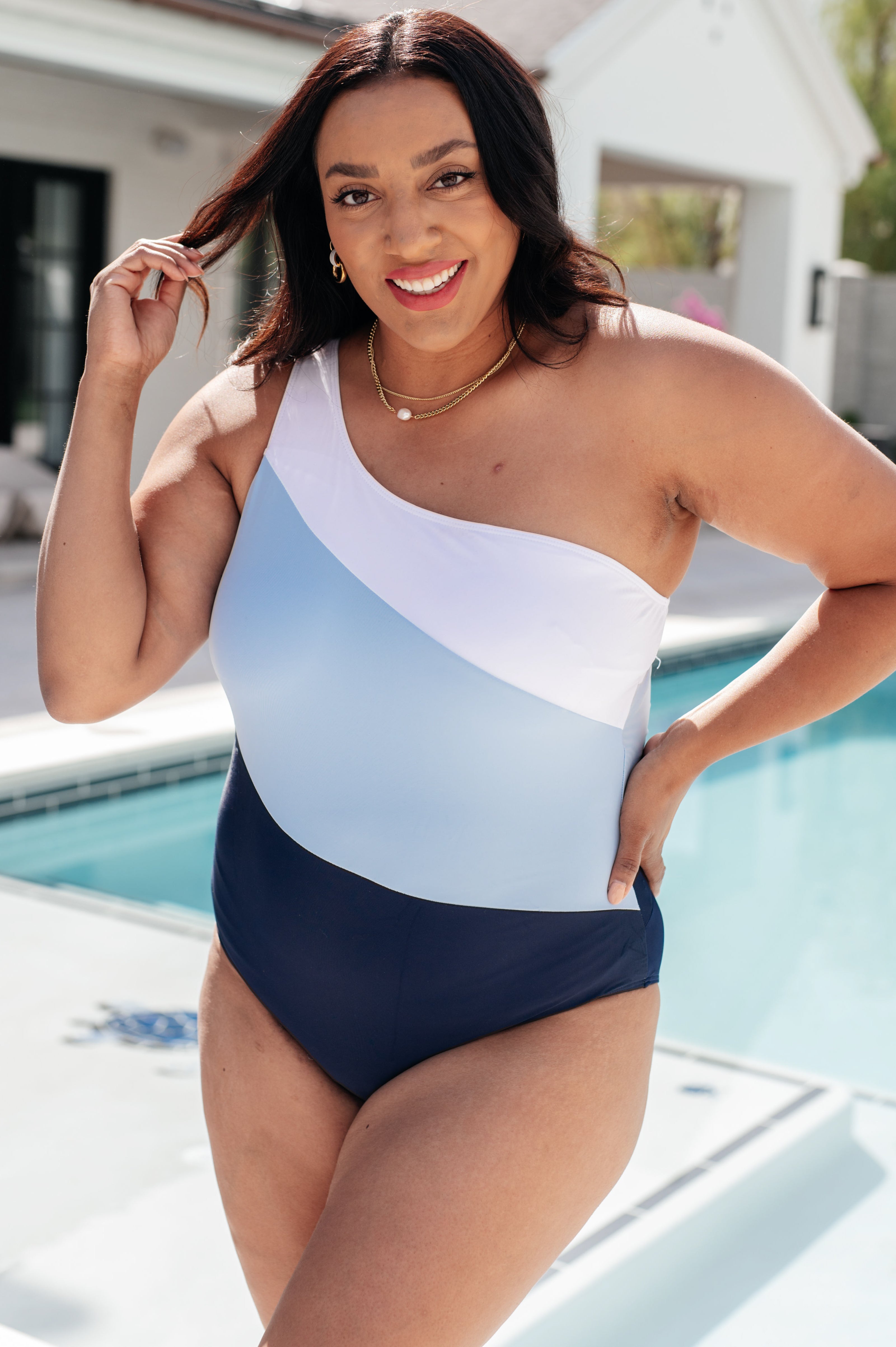 Smiling woman wearing a blue colorblock asymmetrical one-shoulder swimsuit beside a pool outdoors