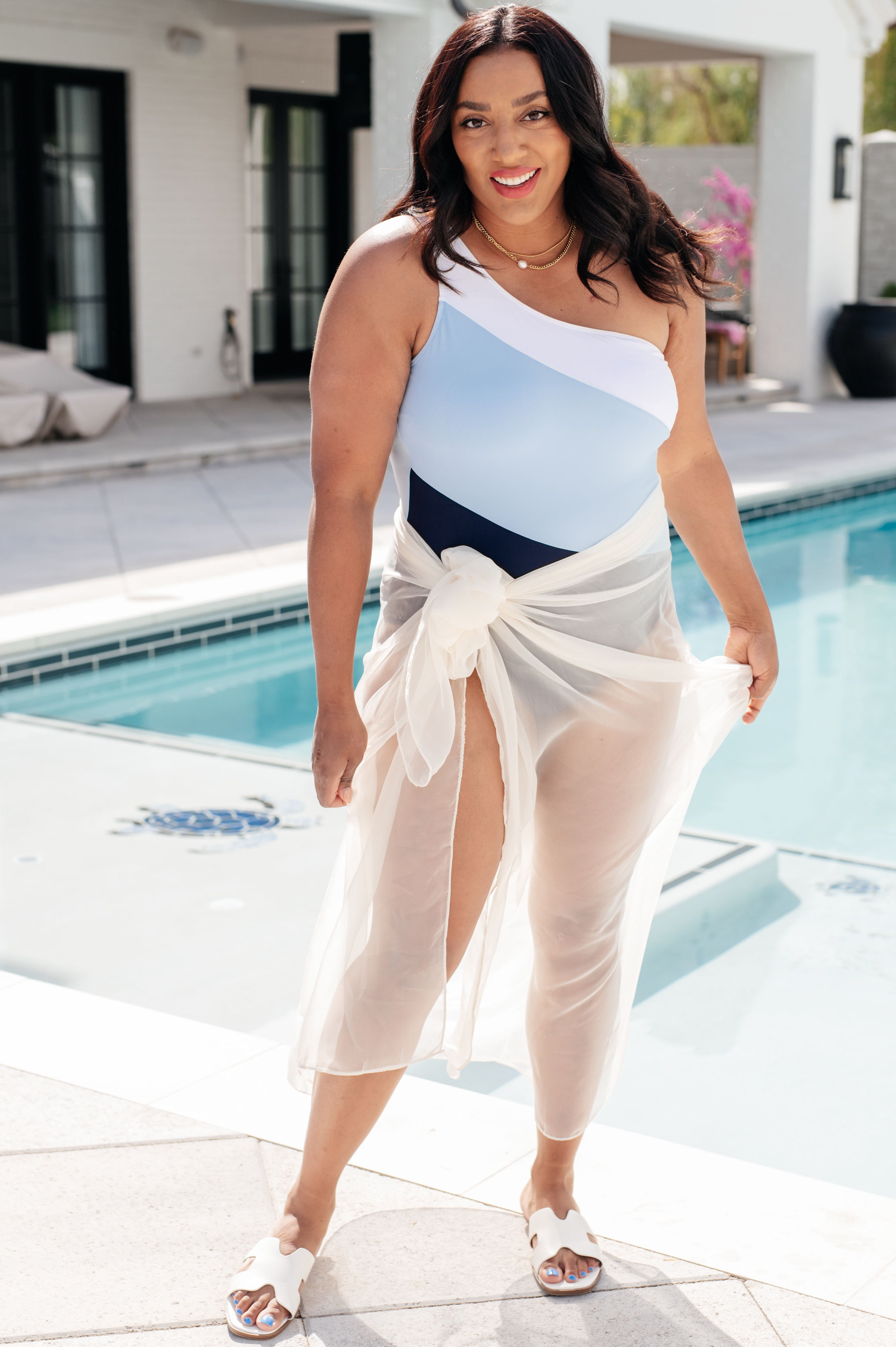 Smiling woman wearing a blue colorblock one-shoulder swimsuit with a sheer white cover-up tied at the waist by a pool