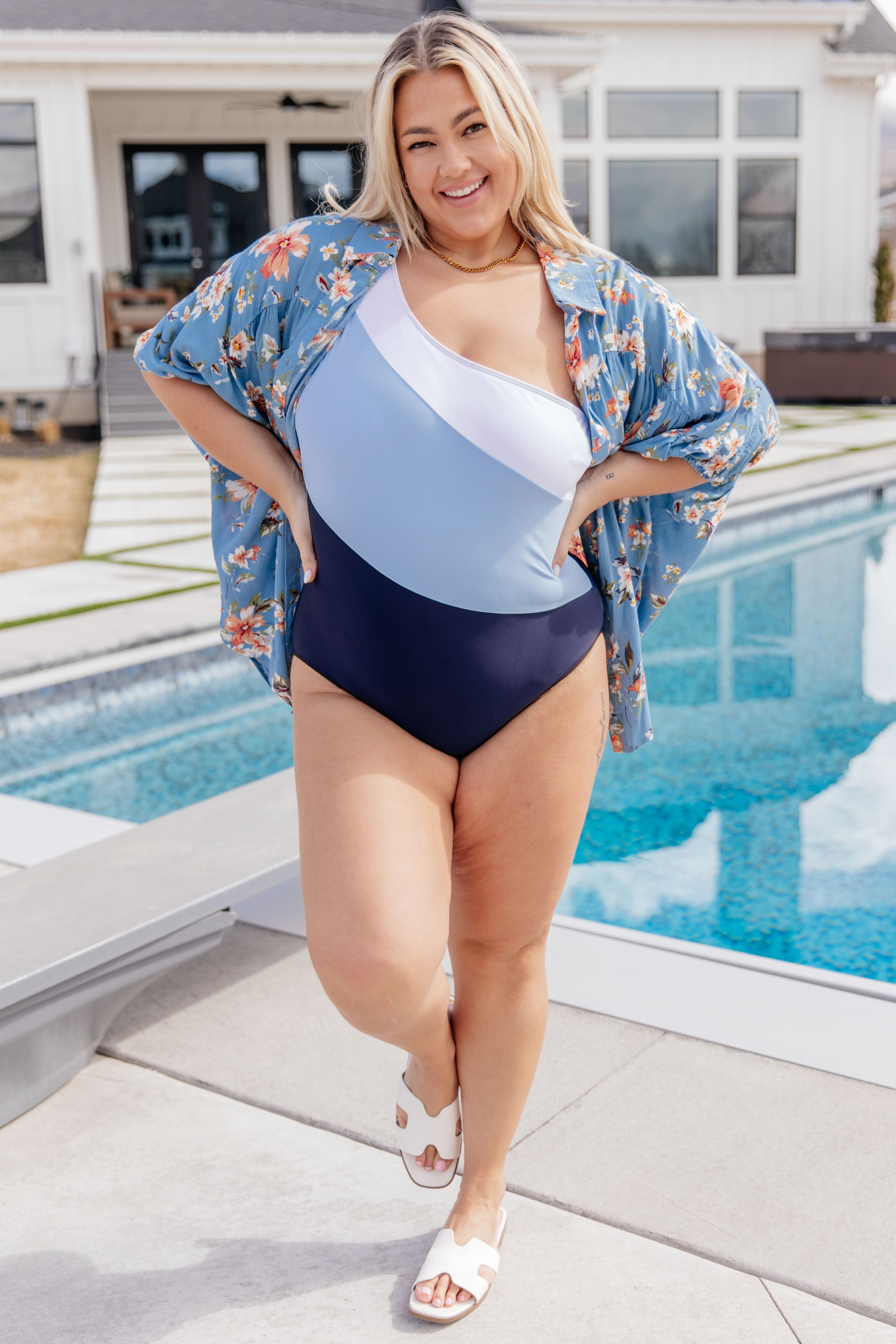 Smiling woman posing by a pool wearing a blue, white, and navy asymmetrical one-piece swimsuit and a floral cover-up