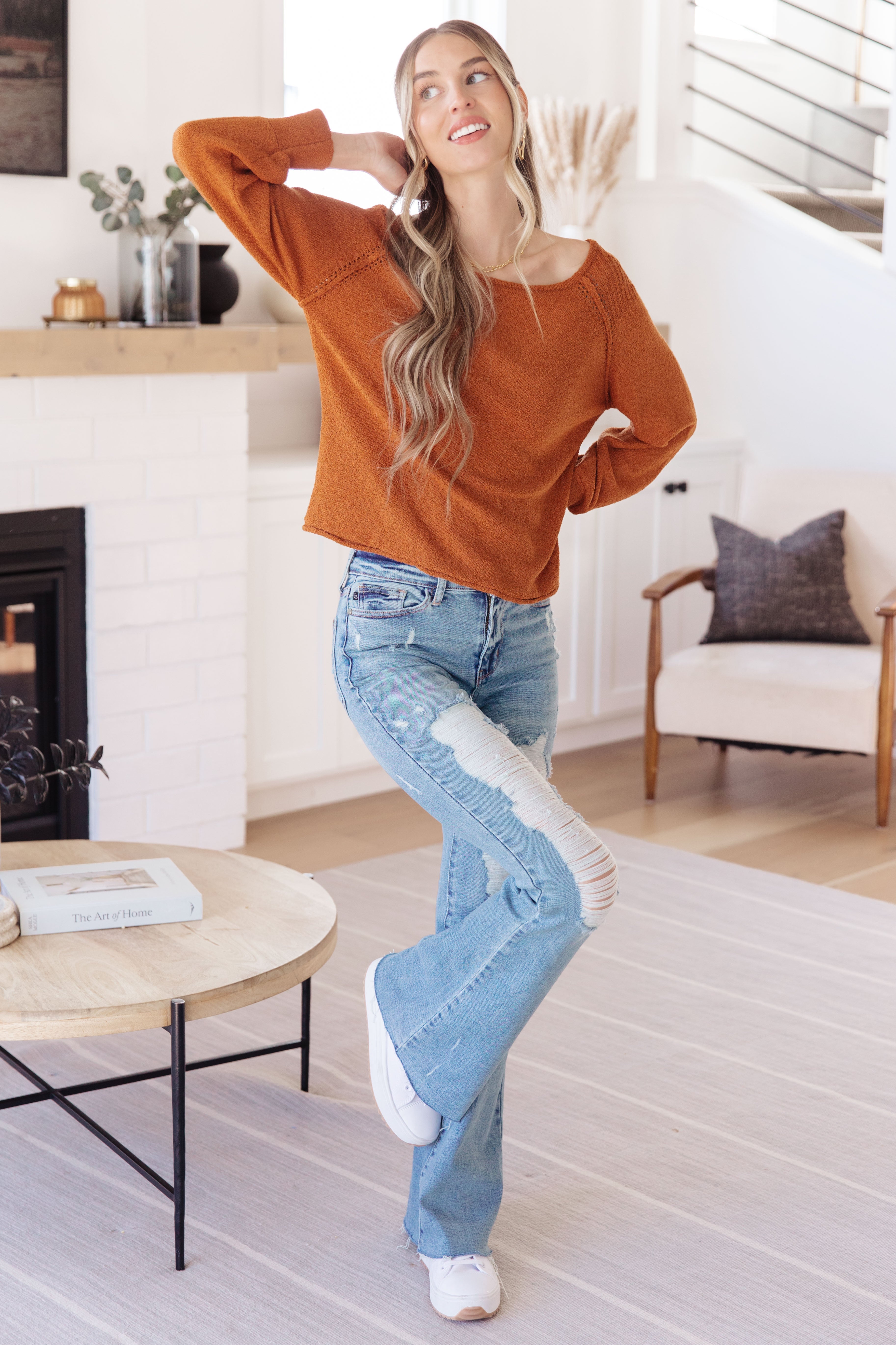 Woman with long blonde hair wearing burnt orange relaxed pullover sweater and ripped light blue jeans indoors