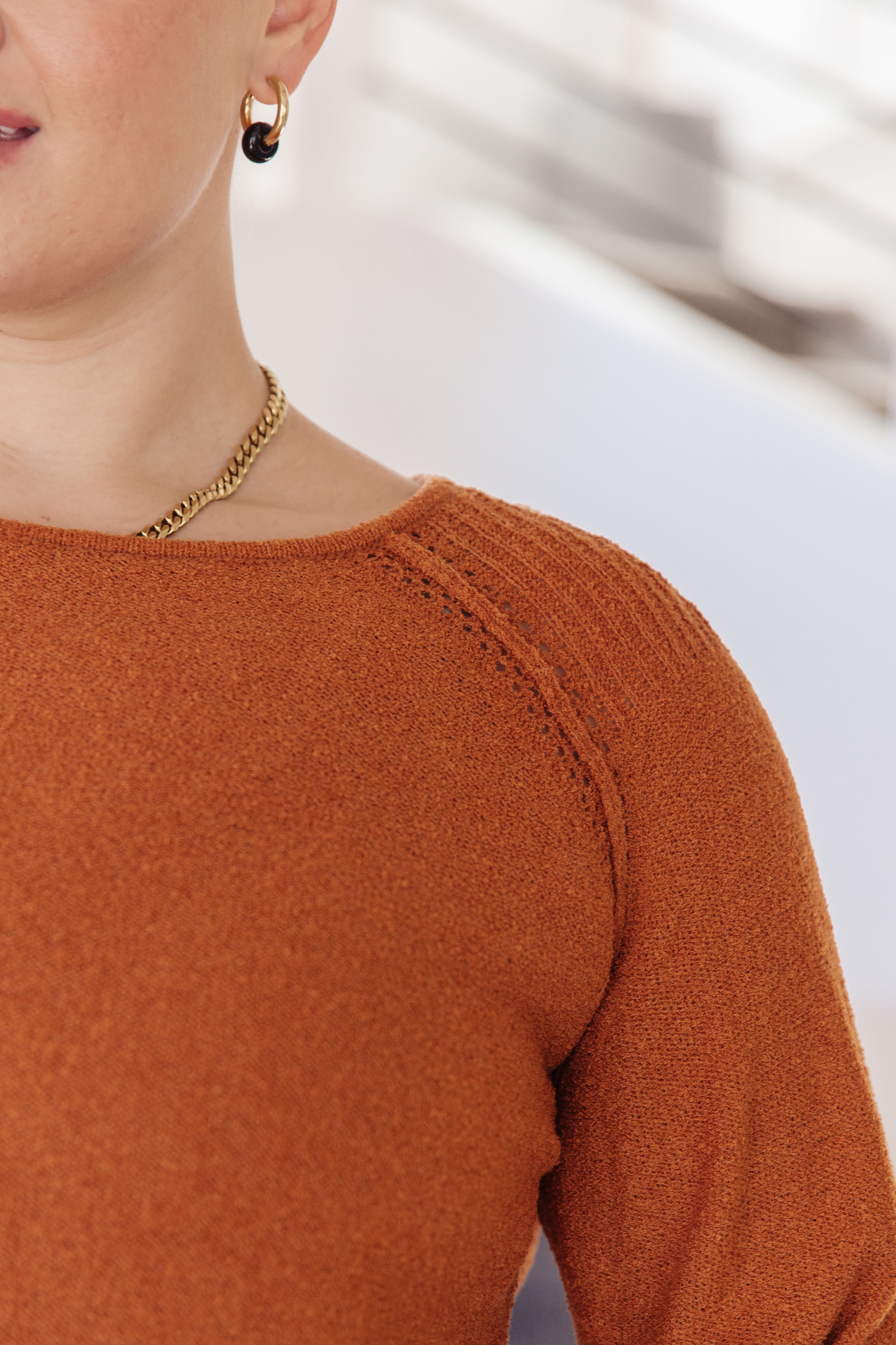 Close-up of person wearing textured rust-colored knitted pullover sweater with gold chain necklace and hoop earring indoors