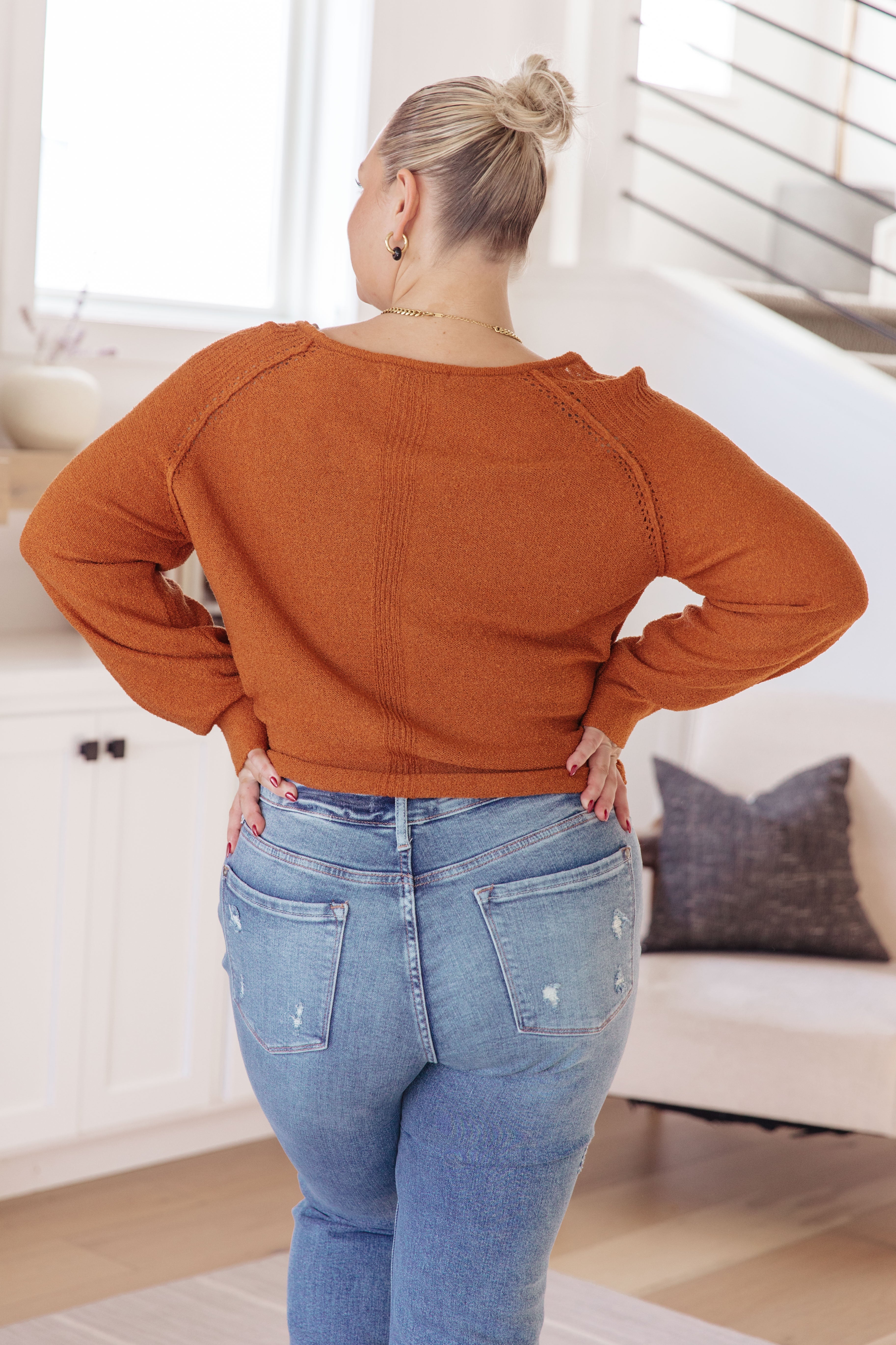 Woman with blonde hair in messy bun wearing rust-colored cropped pullover sweater and distressed blue jeans indoors