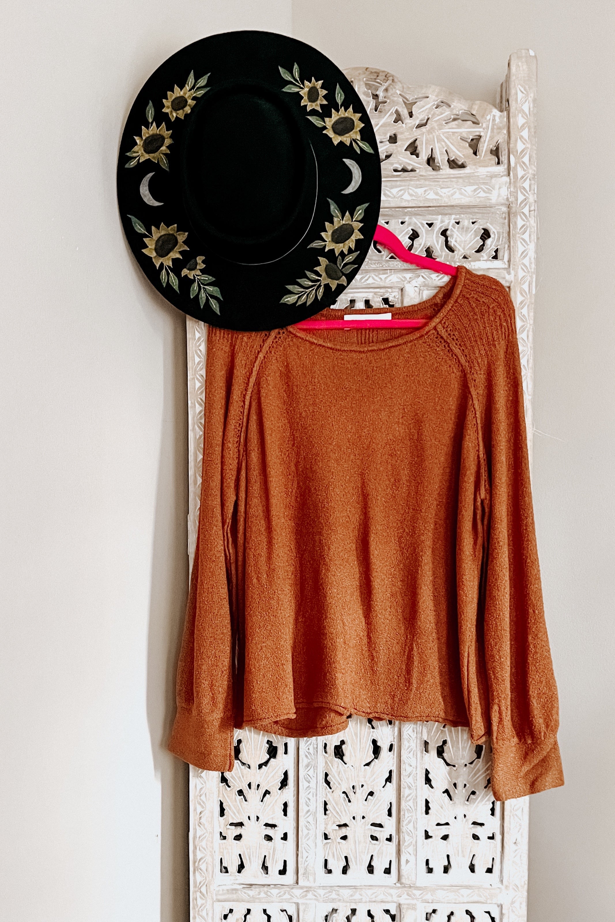 Rust-colored pullover sweater with long sleeves hanging on carved white wooden panel beside black hat with sunflower embroidery