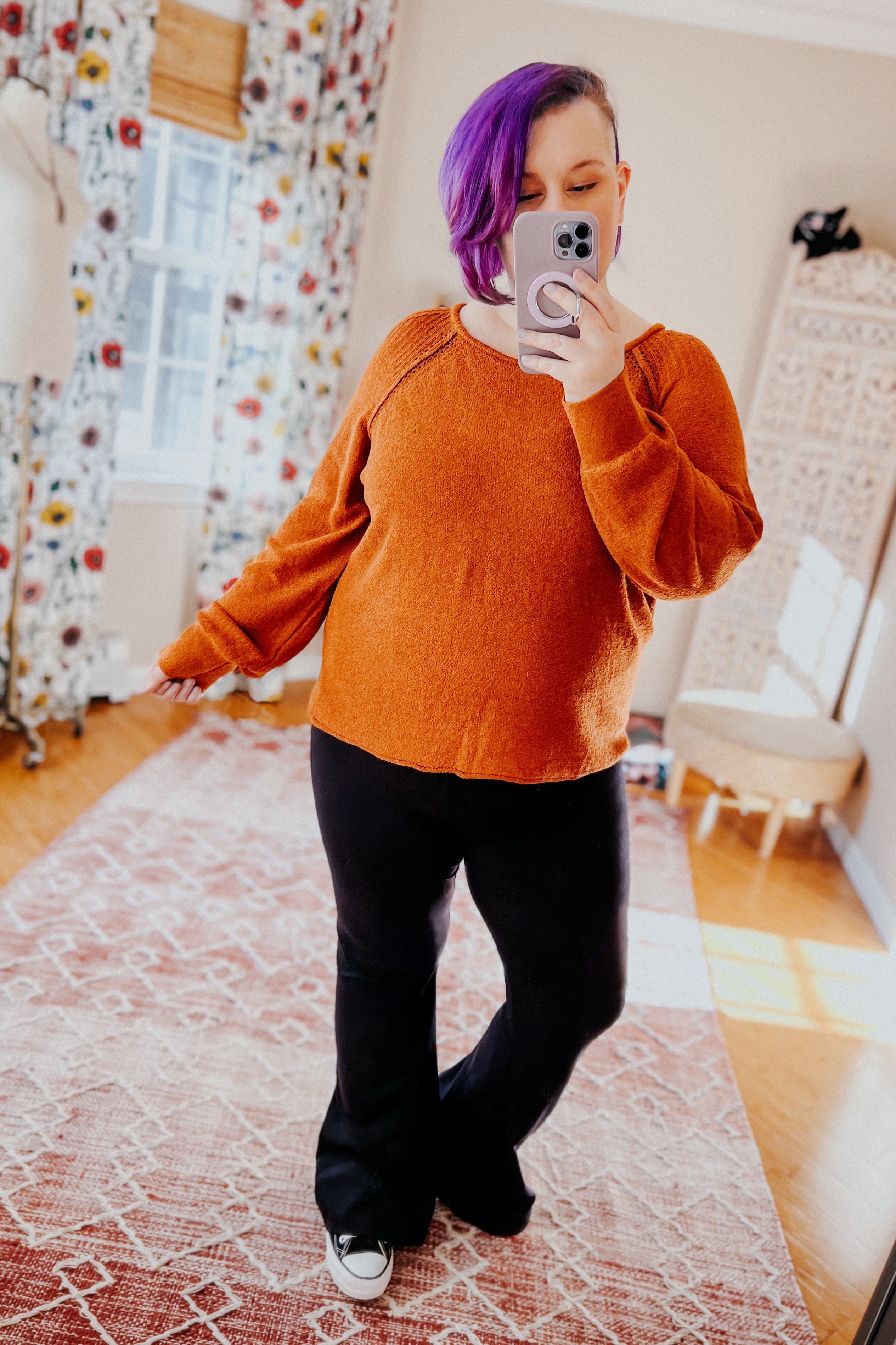 Woman with purple hair wearing rust-colored pullover sweater and black pants taking mirror selfie in cozy room