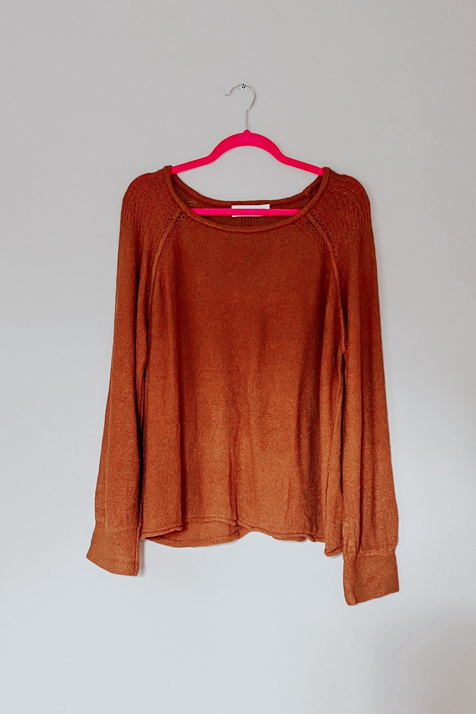 Rust-colored loose pullover sweater with long sleeves hanging on a bright pink hanger against plain wall
