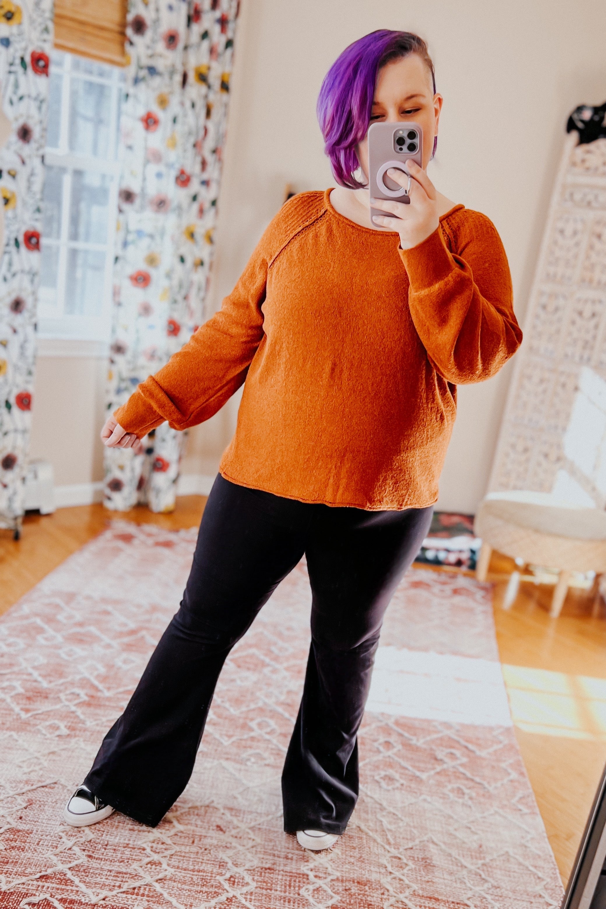 Woman with short purple hair wearing a rust-colored pullover sweater and black flared pants taking a mirror selfie indoors