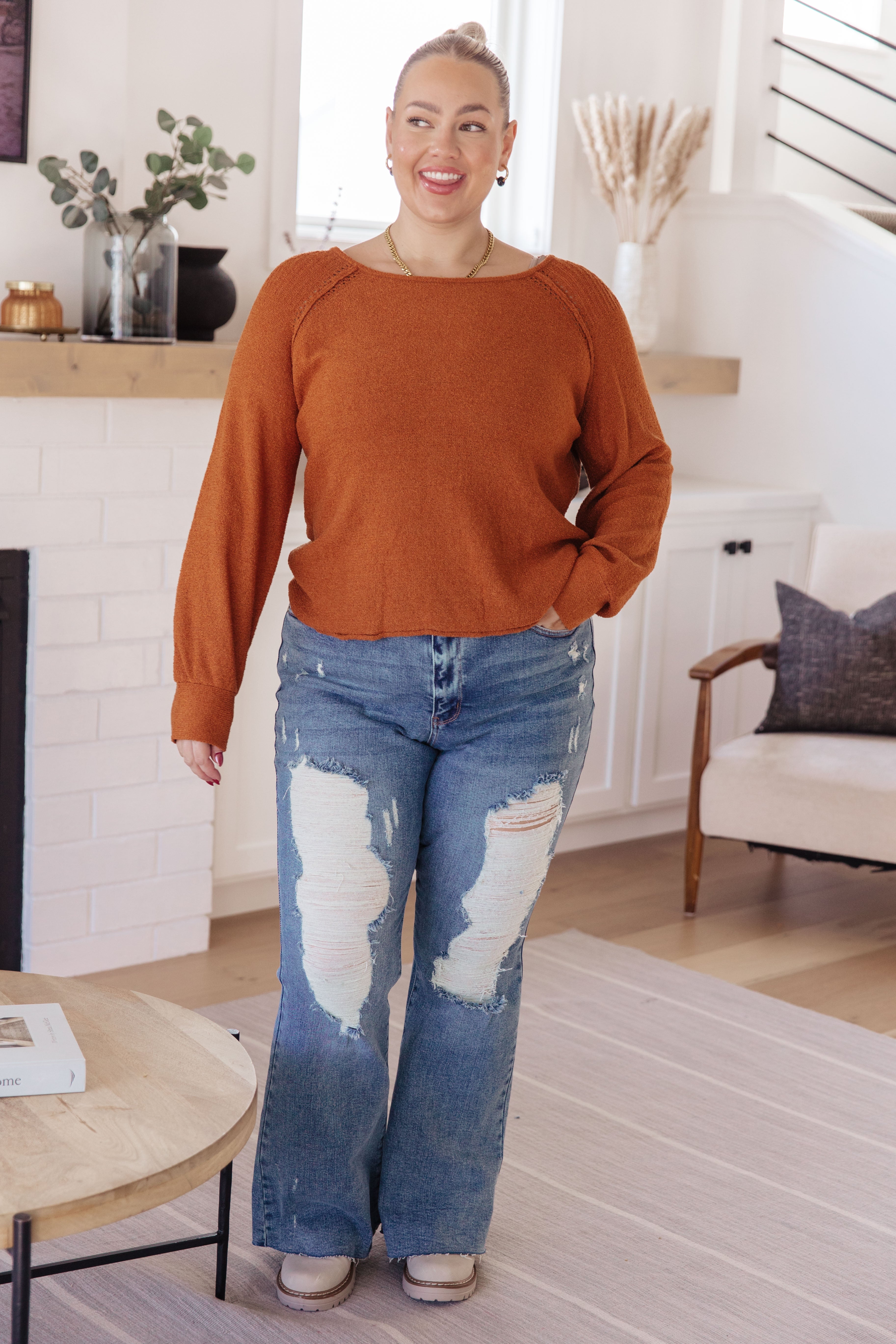 Woman with hair in a bun wearing burnt orange pullover sweater and heavily distressed wide-leg jeans indoors