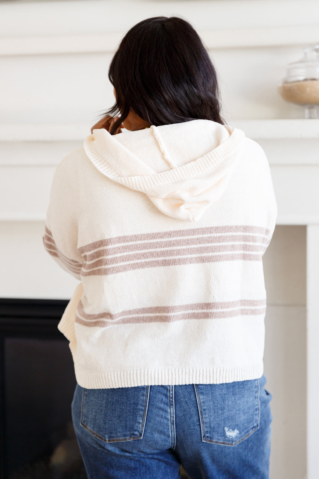 Back view of a woman wearing a cream hooded sweater with brown stripes and blue jeans indoors