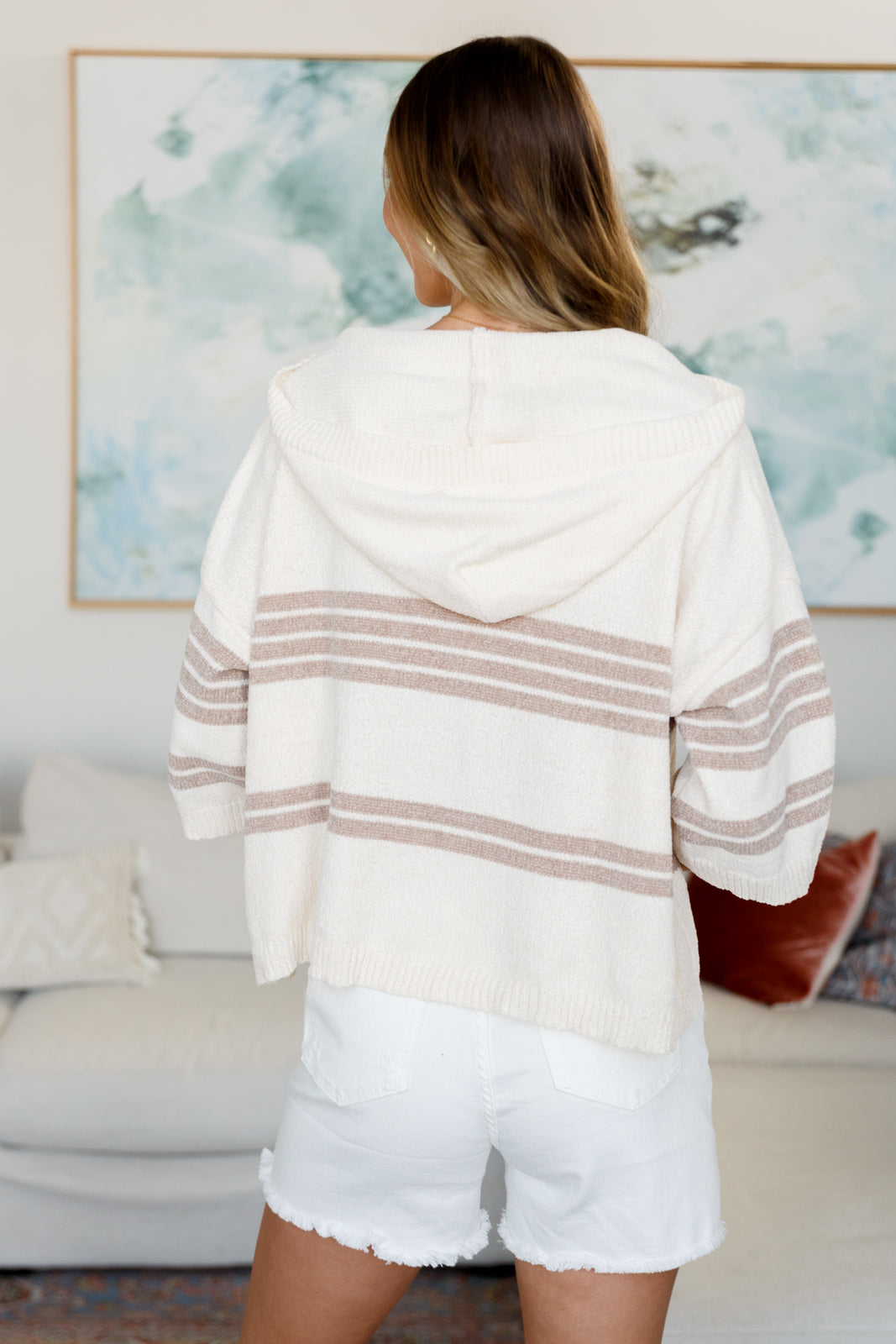 Back view of woman wearing a cream striped hooded sweater with white distressed shorts indoors