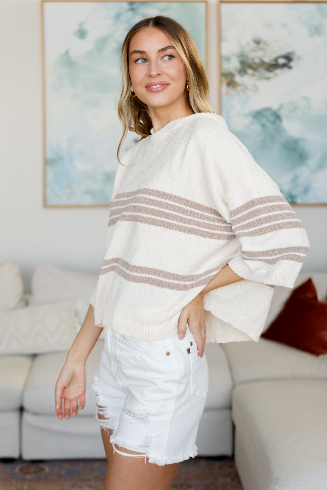 Woman modeling a cream striped hoodie with brown accents and distressed white denim shorts indoors