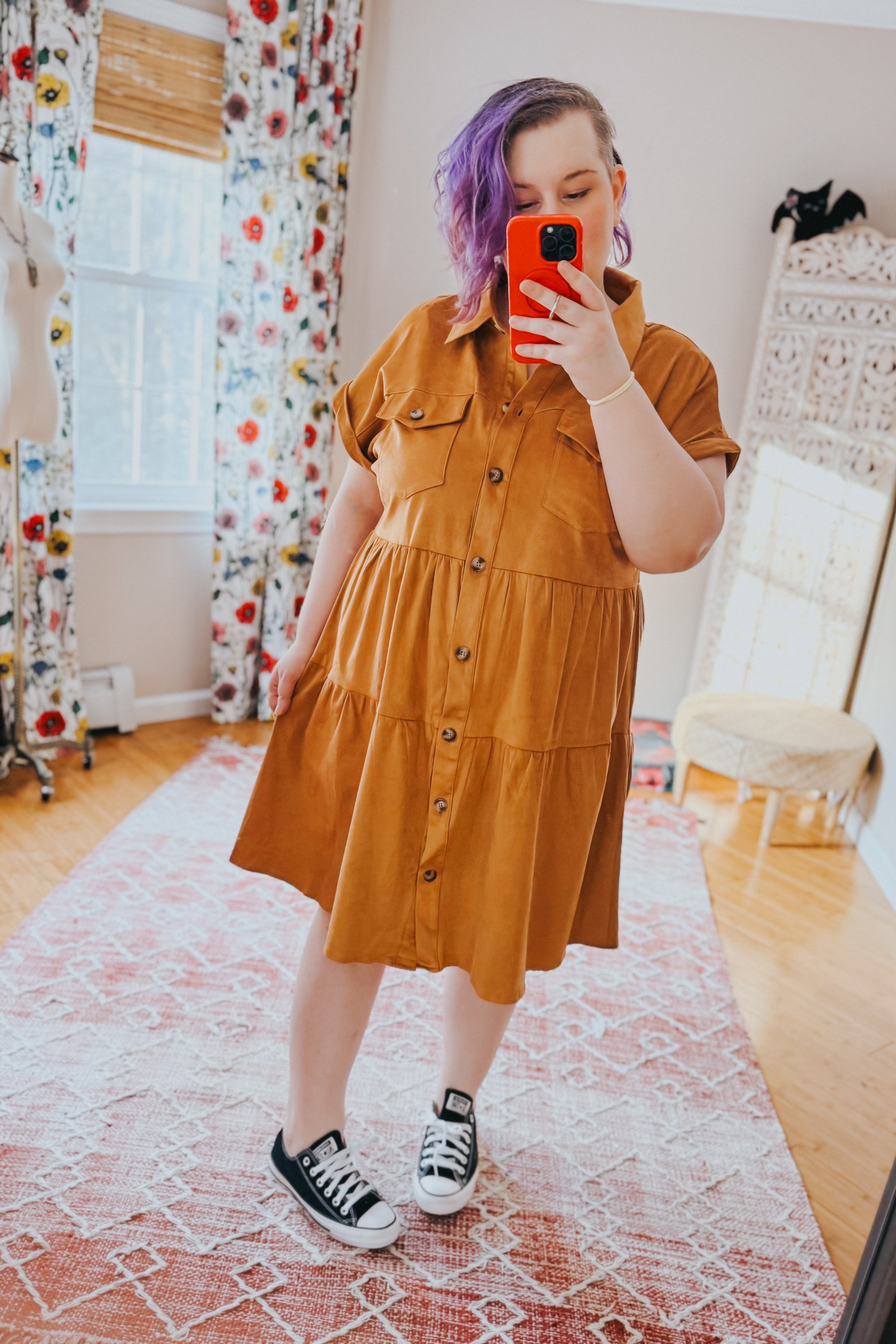 Woman with purple hair wearing a tan faux suede button-up shirt dress and black sneakers taking a mirror selfie indoors