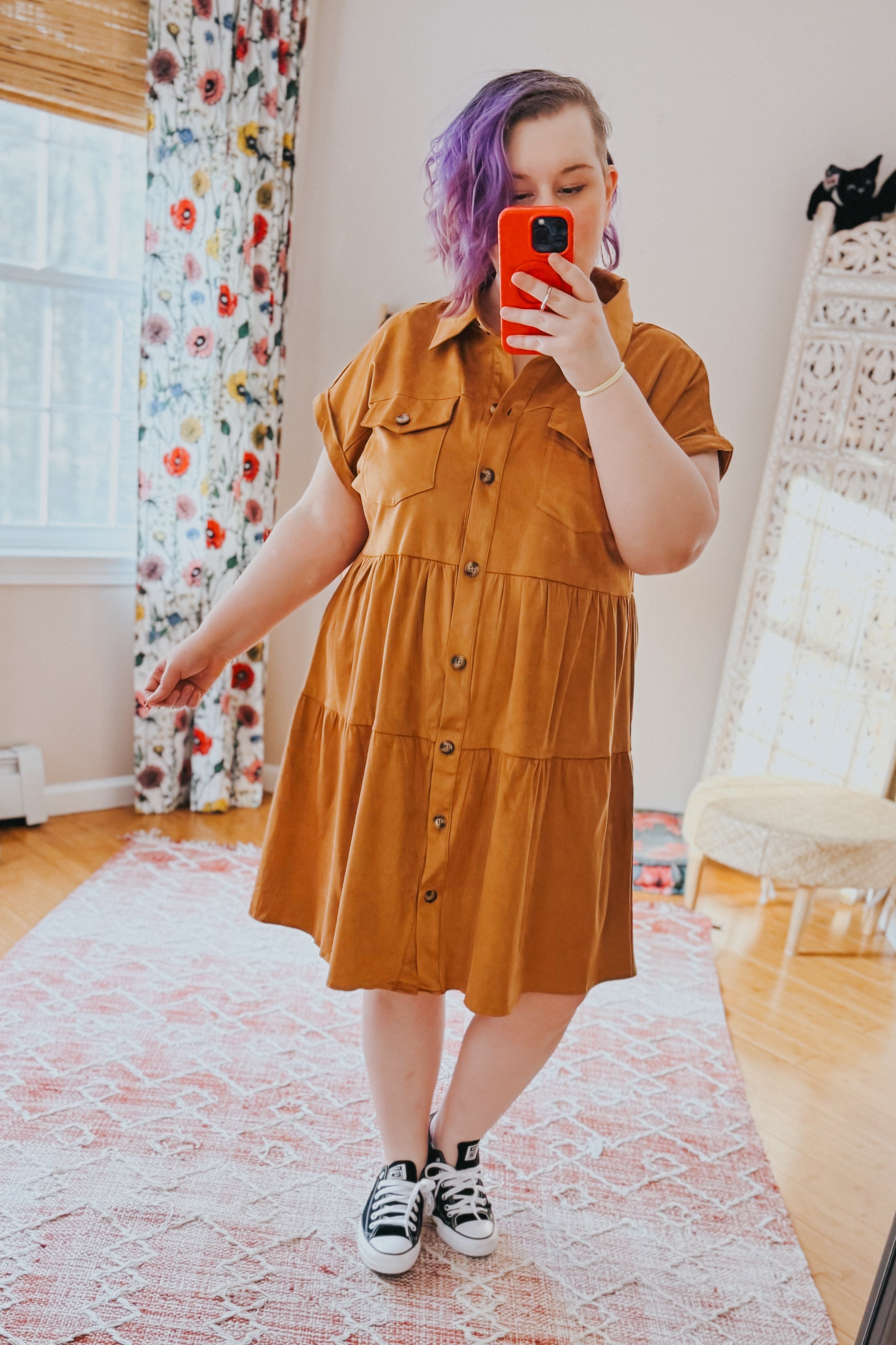 Woman with purple hair wearing a tan faux suede button-up shirt dress and black sneakers taking a mirror selfie indoors