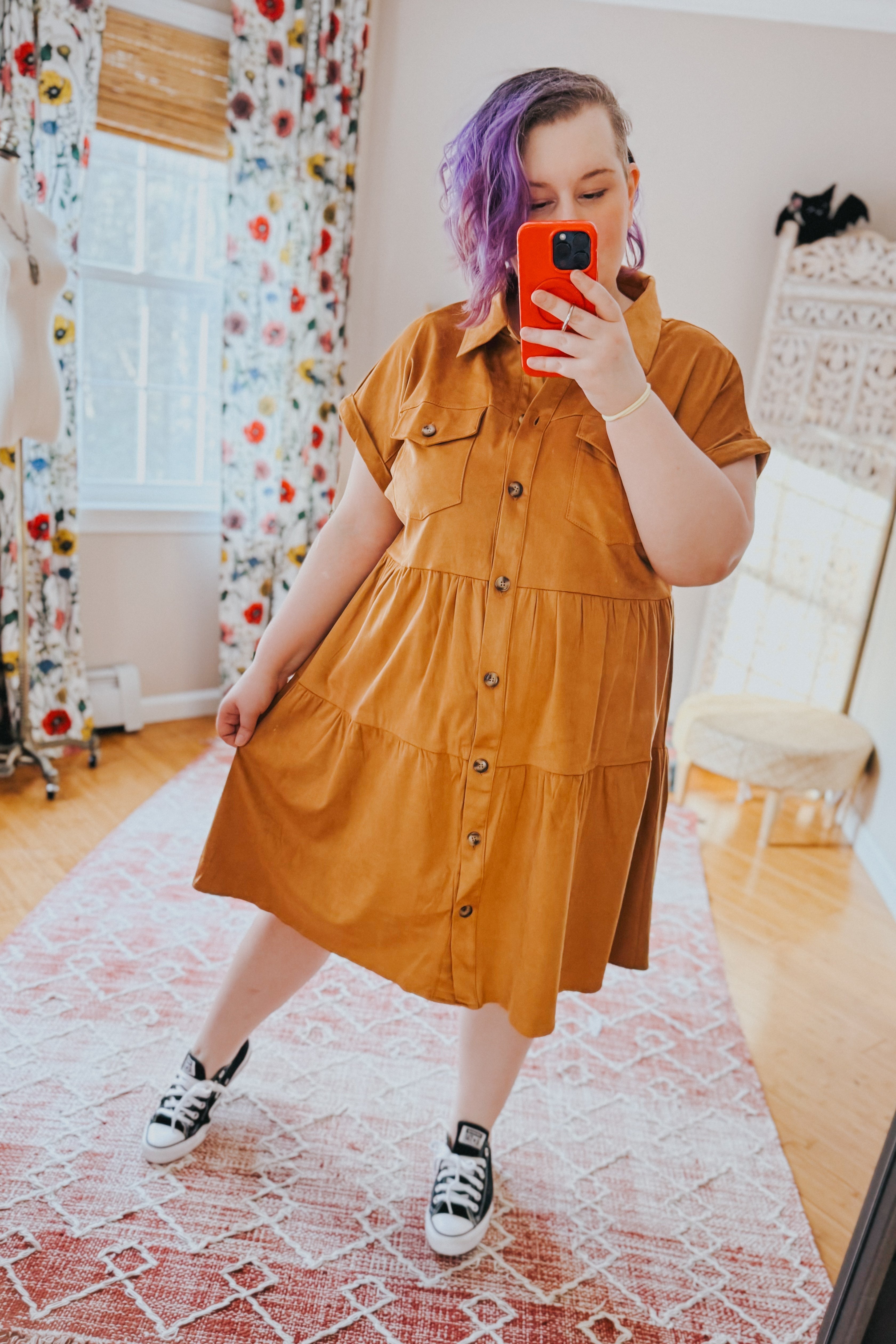 Woman wearing a mustard faux suede button-up shirt dress with pockets and black sneakers taking a mirror selfie indoors