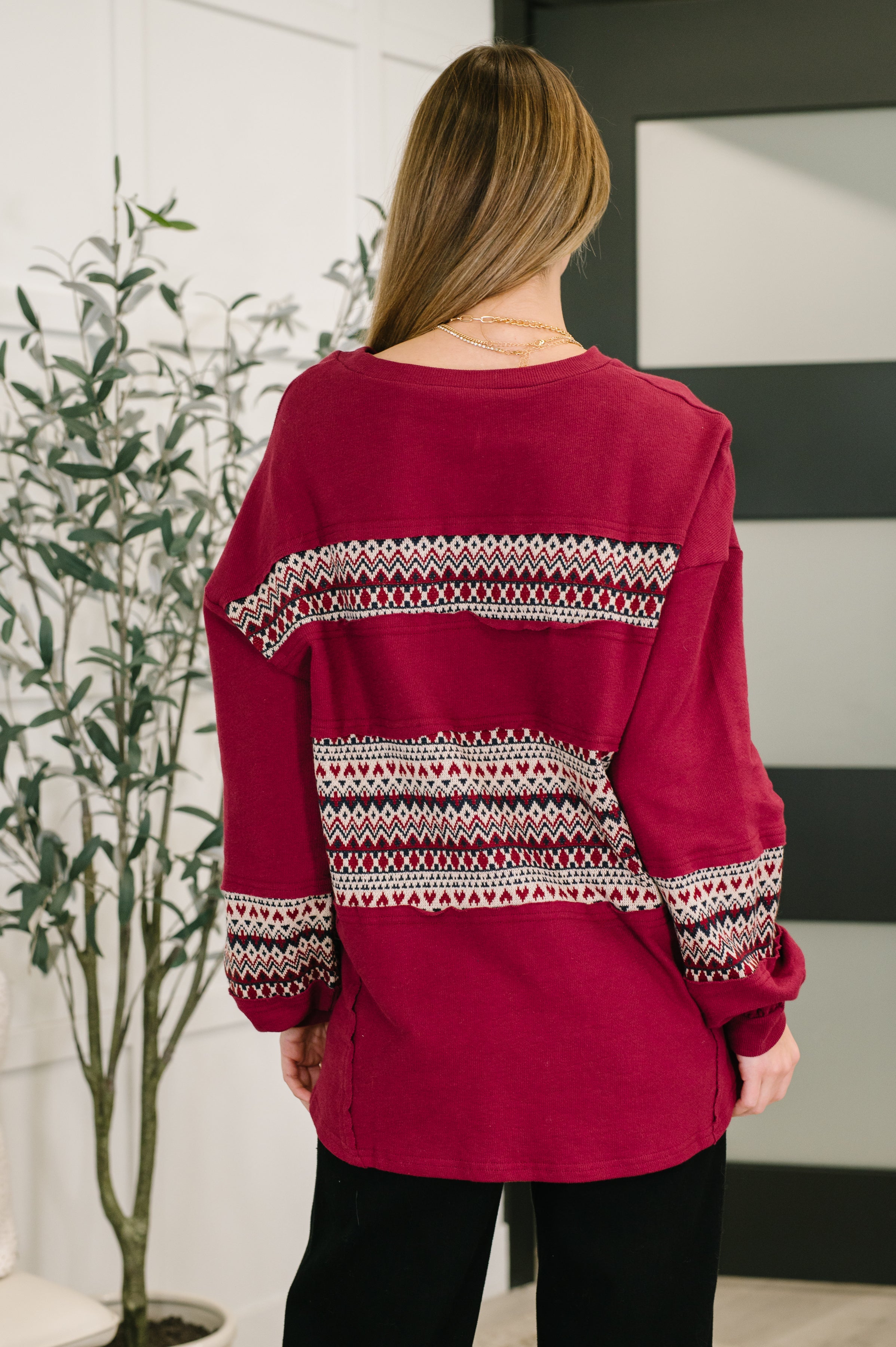 Woman wearing a long sleeve patterned red sweater, a gold necklace and black pants standing in an indoor setting.