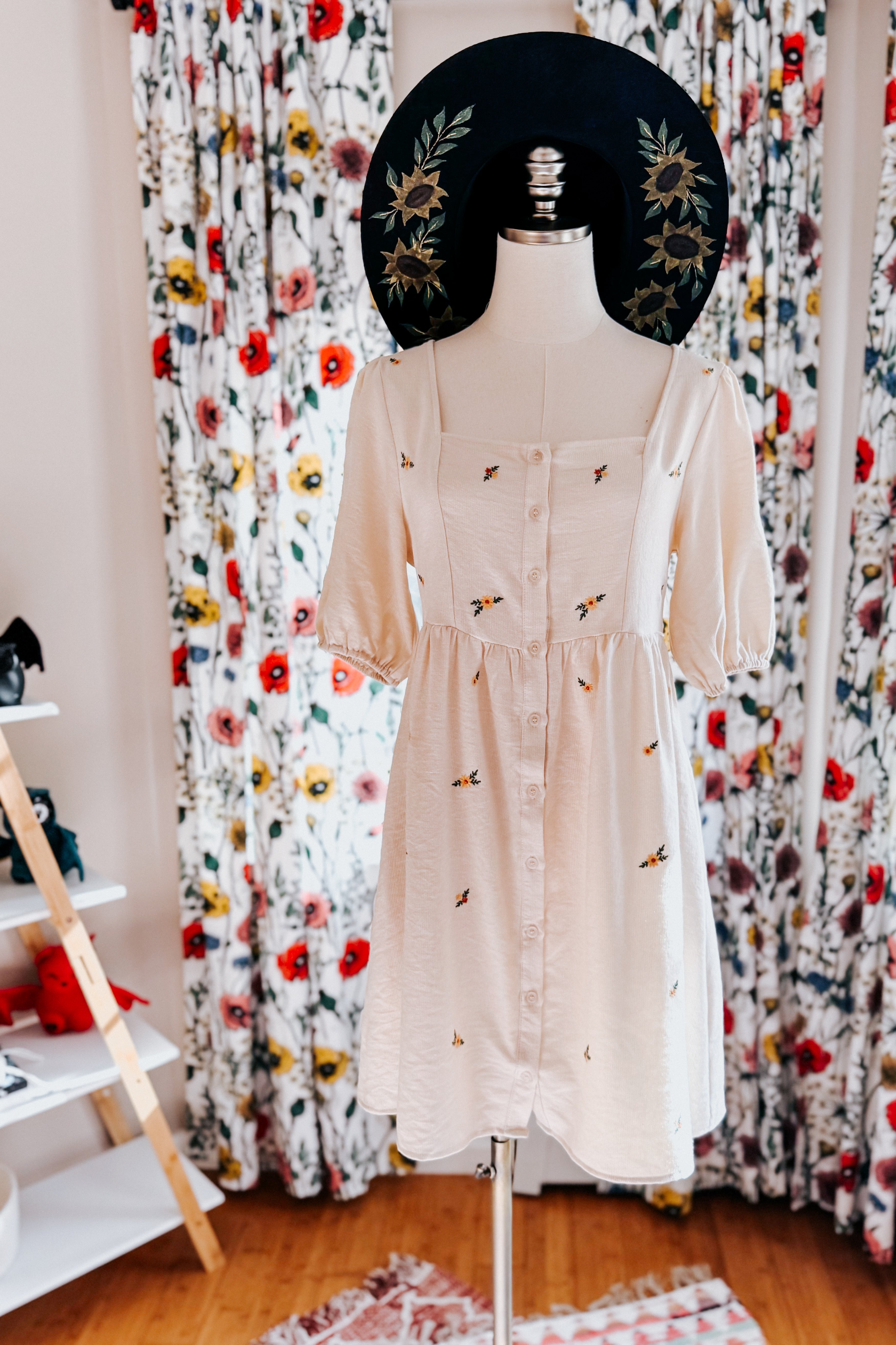 Cream knee-length dress with button front, puff sleeves, and small colorful floral embroidery displayed on mannequin with black hat