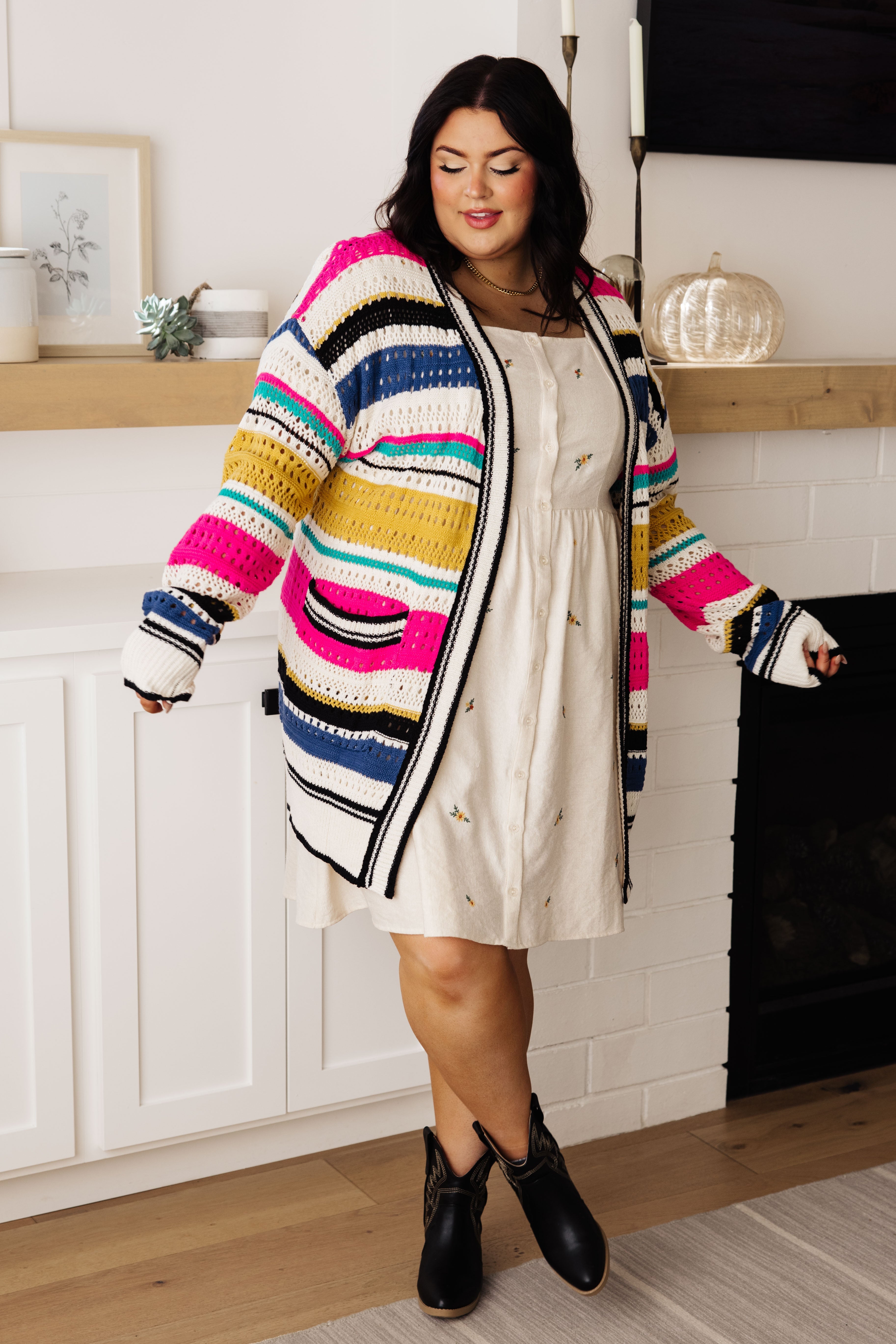 Plus size model wearing cream embroidered dress with square neckline and button front paired with colorful striped cardigan indoors