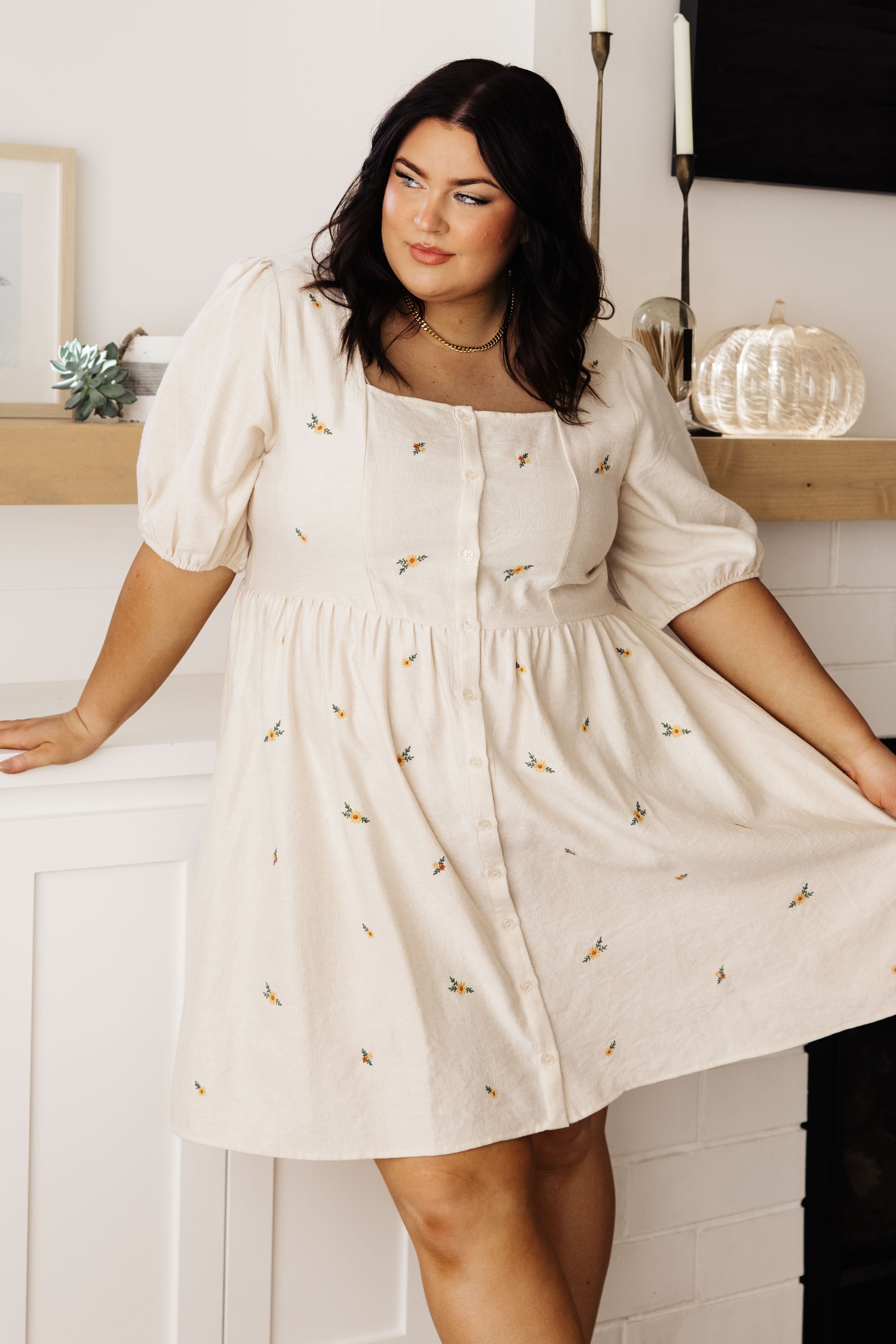Plus size model wearing cream dress with square neckline, puff sleeves, button front, and scattered floral embroidery indoors