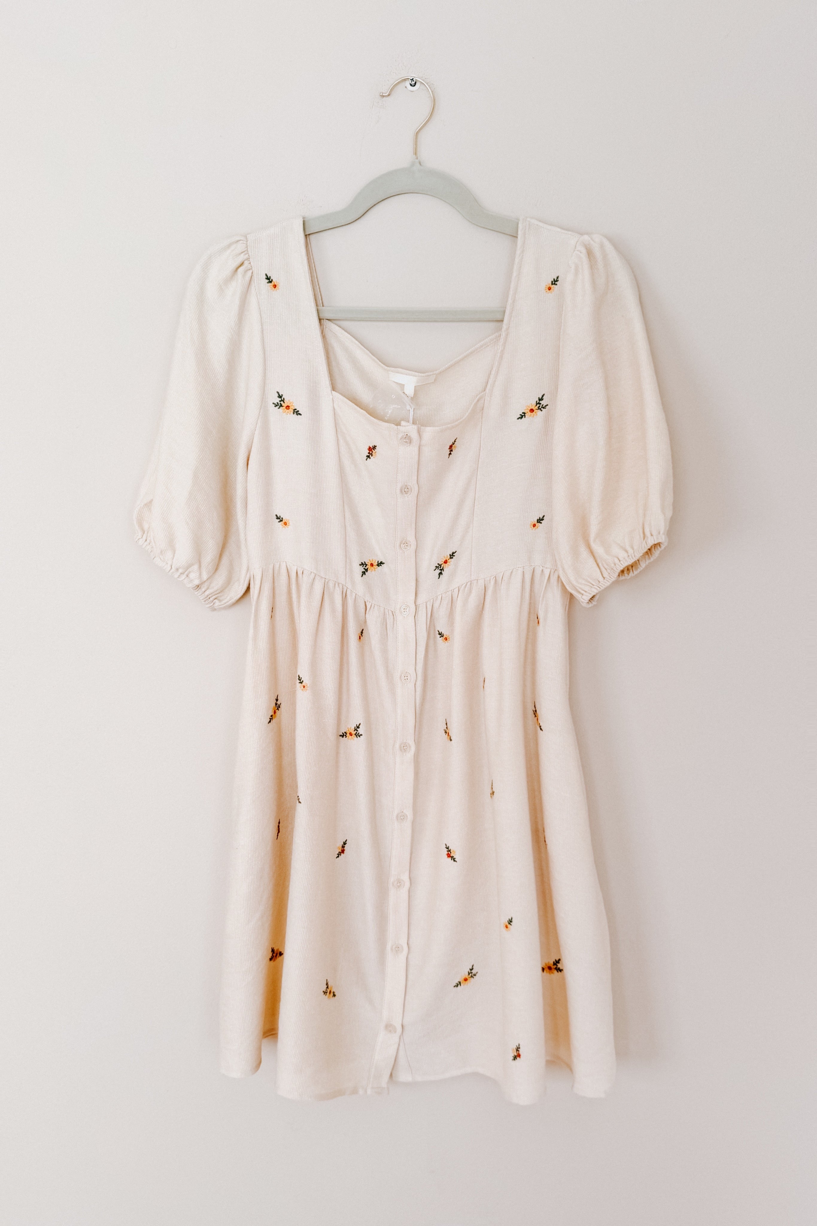Cream button-front dress with puff sleeves and delicate orange floral embroidery hanging on gray hanger against white wall