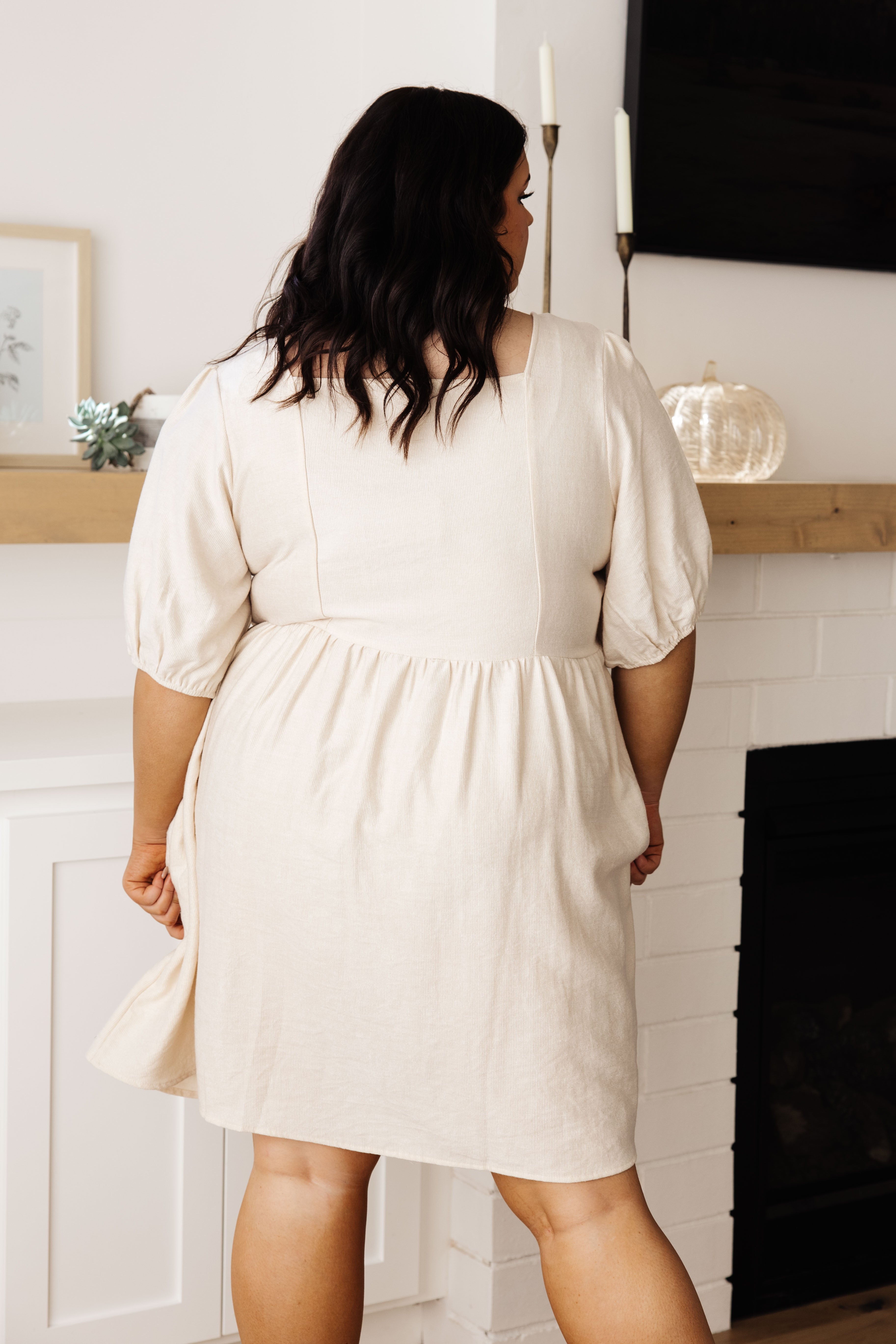 Plus size model wearing cream embroidered dress with puff sleeves and square neckline, shown from back indoors