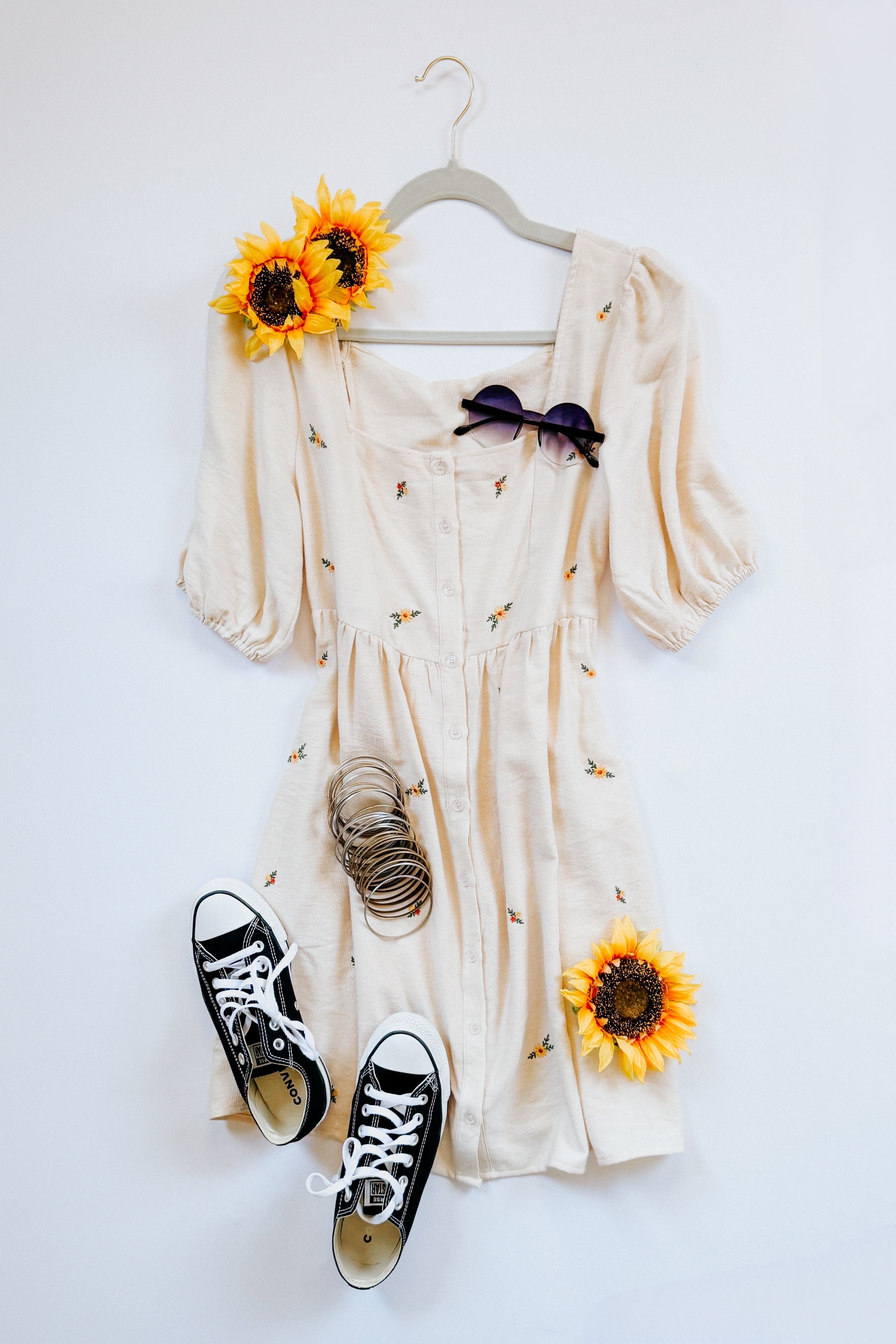 Cream embroidered dress with puff sleeves and buttons displayed with black sneakers, sunglasses, bracelets, and sunflowers