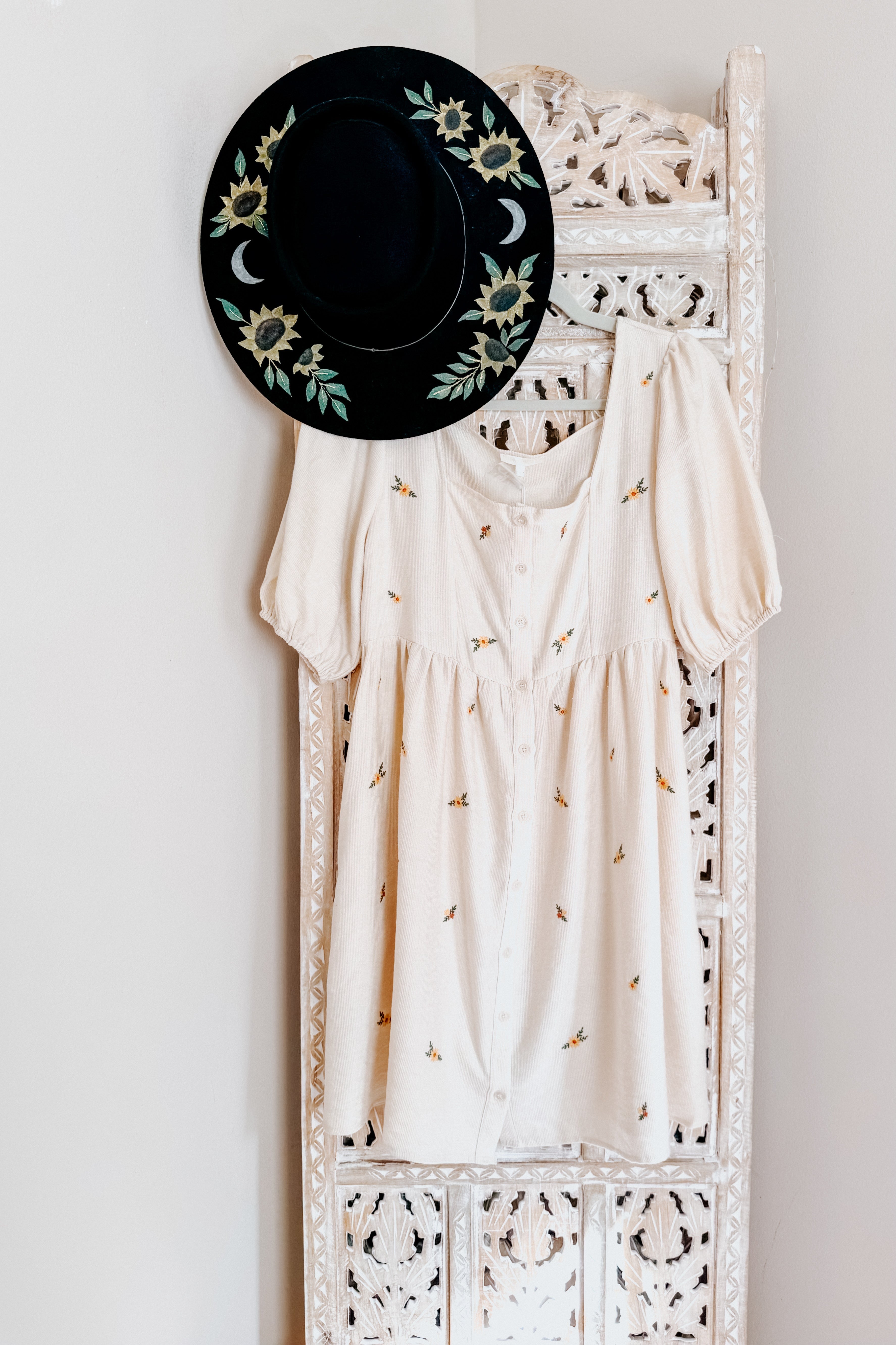 Cream dress with small floral embroidery and puff sleeves hanging on carved white wooden panel next to black floral hat