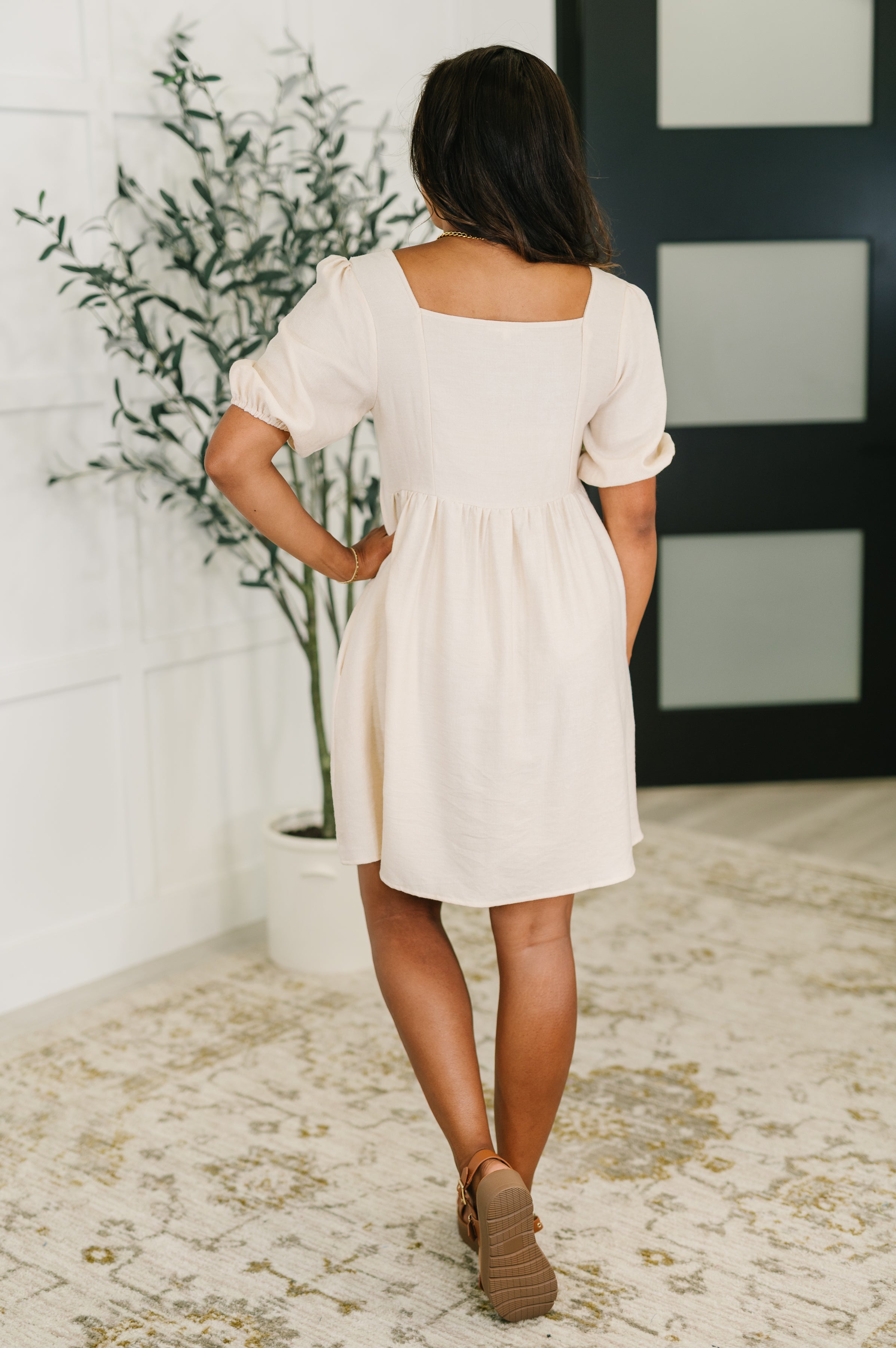 Woman seen from back wearing cream knee-length dress with puff sleeves and square neckline indoors