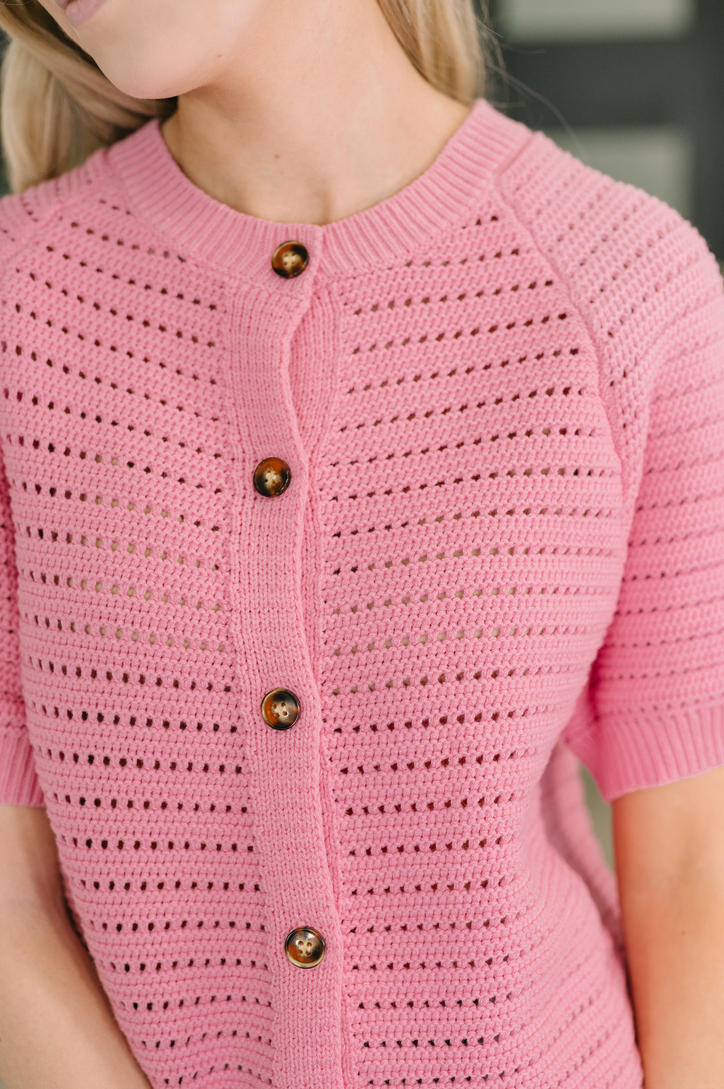 Close view of a woman wearing a pink knit button down sweater with a ribbed neckline short sleeves and wood tone buttons