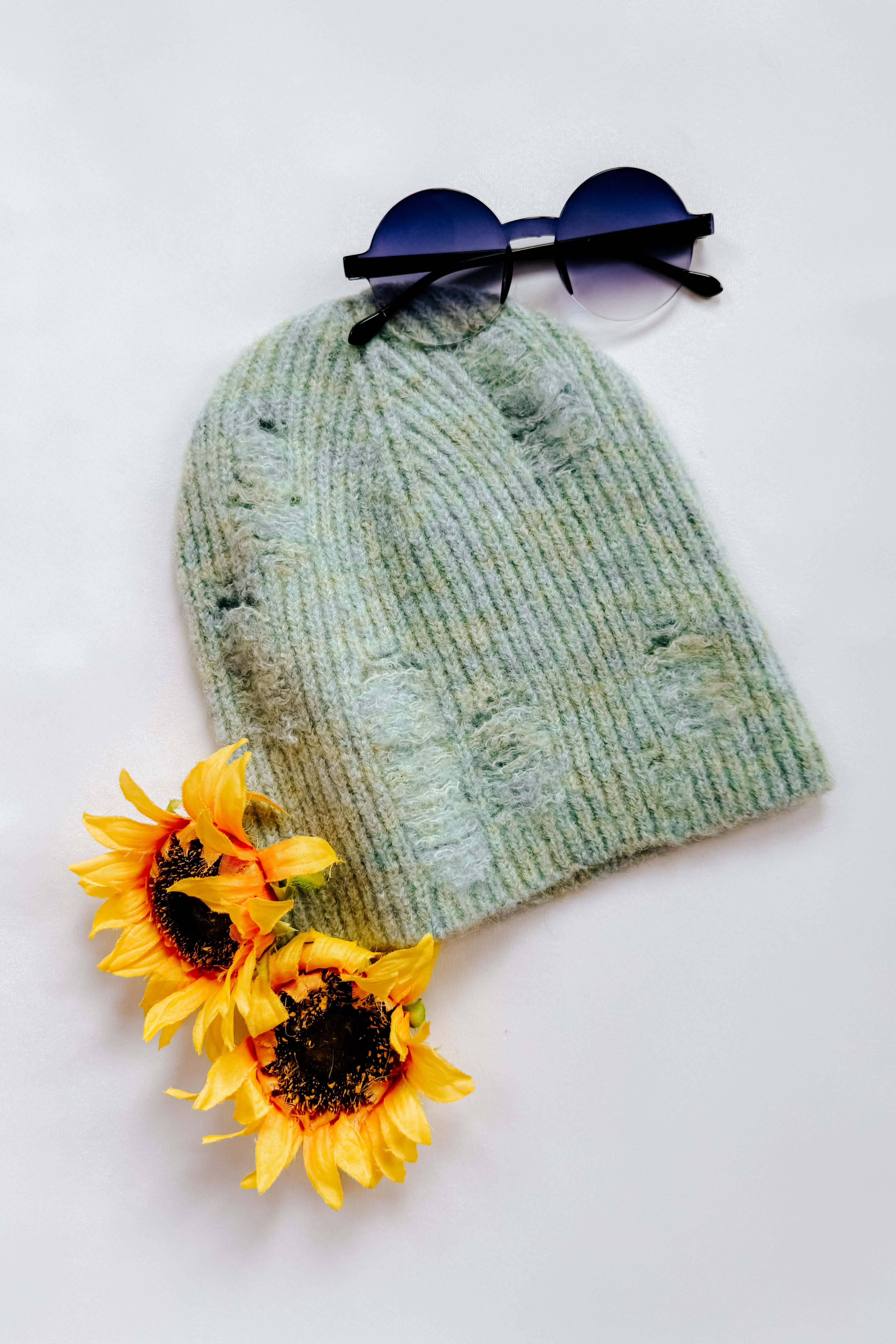 Soft mint green distressed ribbed knit beanie paired with round ombre sunglasses and two vibrant sunflowers on white background