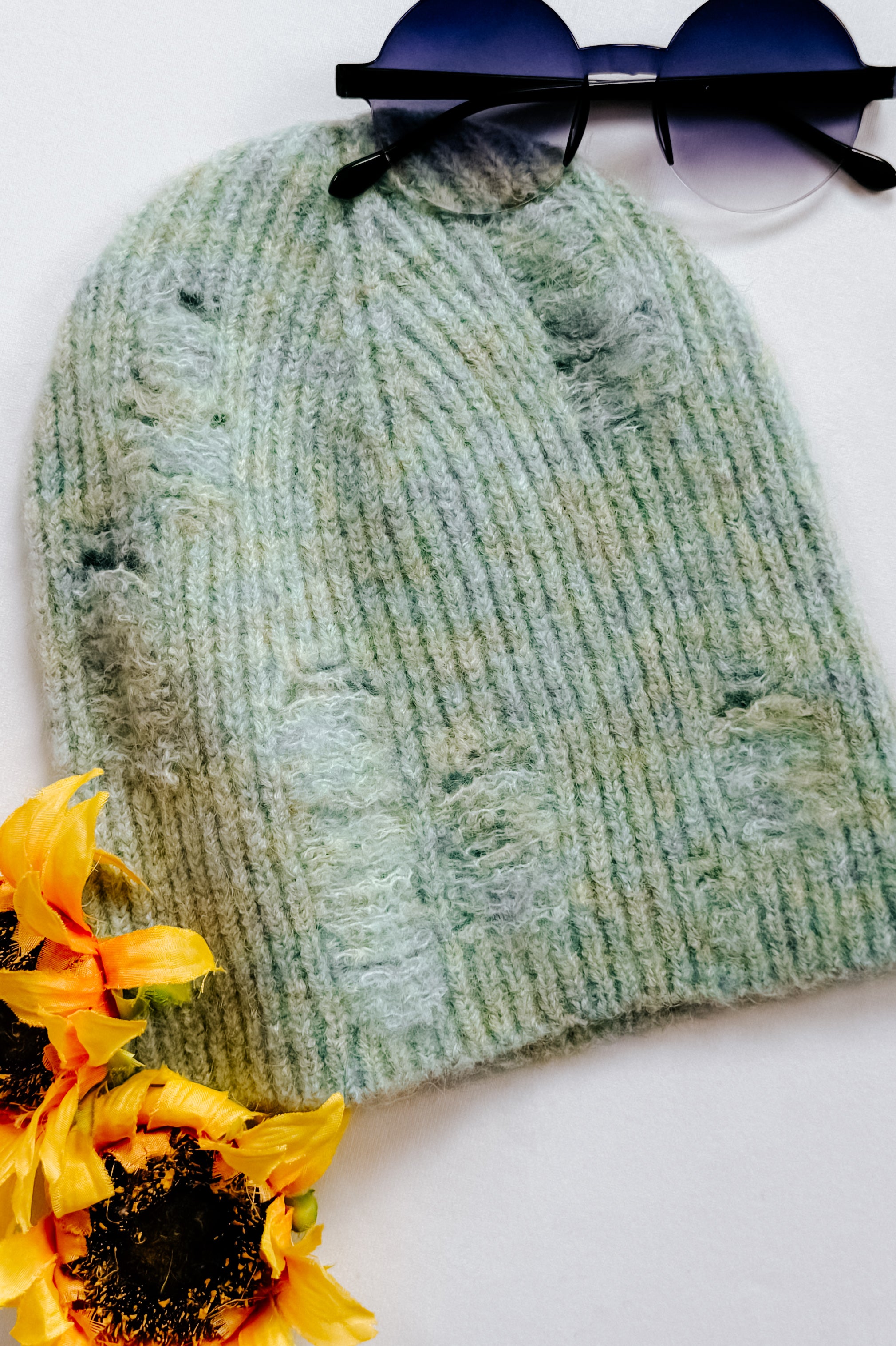 Soft mint green knit beanie with distressed details styled with round sunglasses and sunflowers on white background