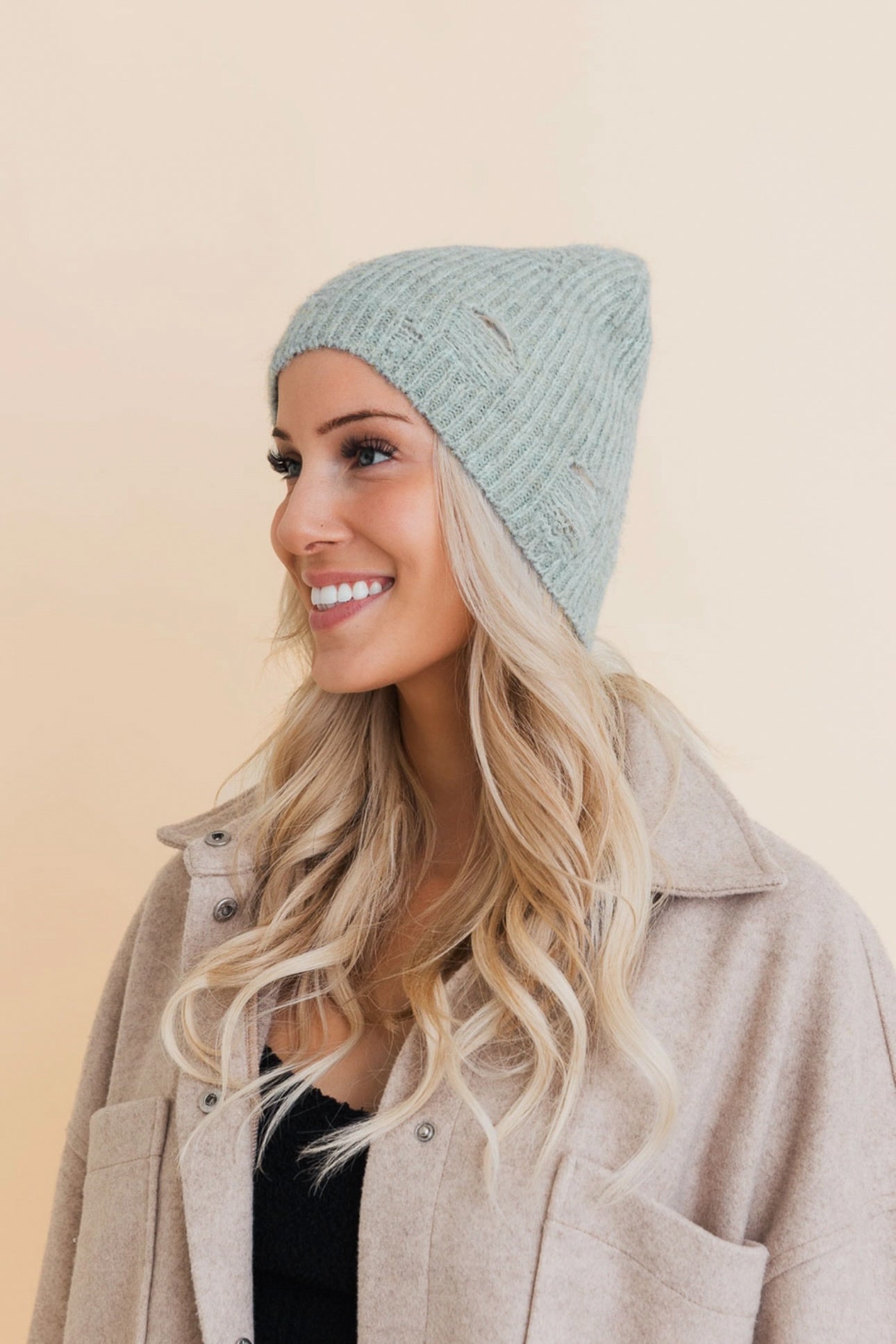 Woman smiling wearing a soft mint ribbed knit beanie with small distressed holes and beige wool jacket indoors