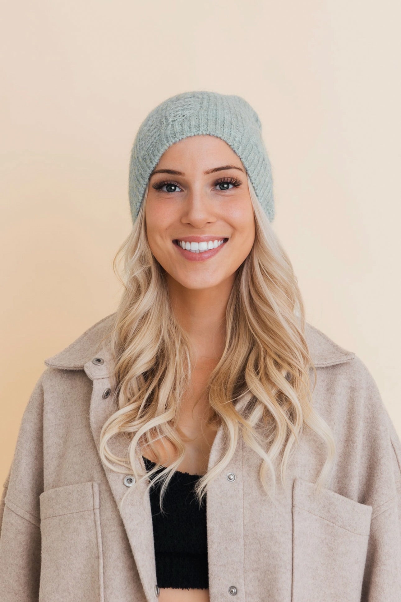 Smiling woman wearing a soft mint knit distressed beanie and beige button-up jacket indoors against neutral background