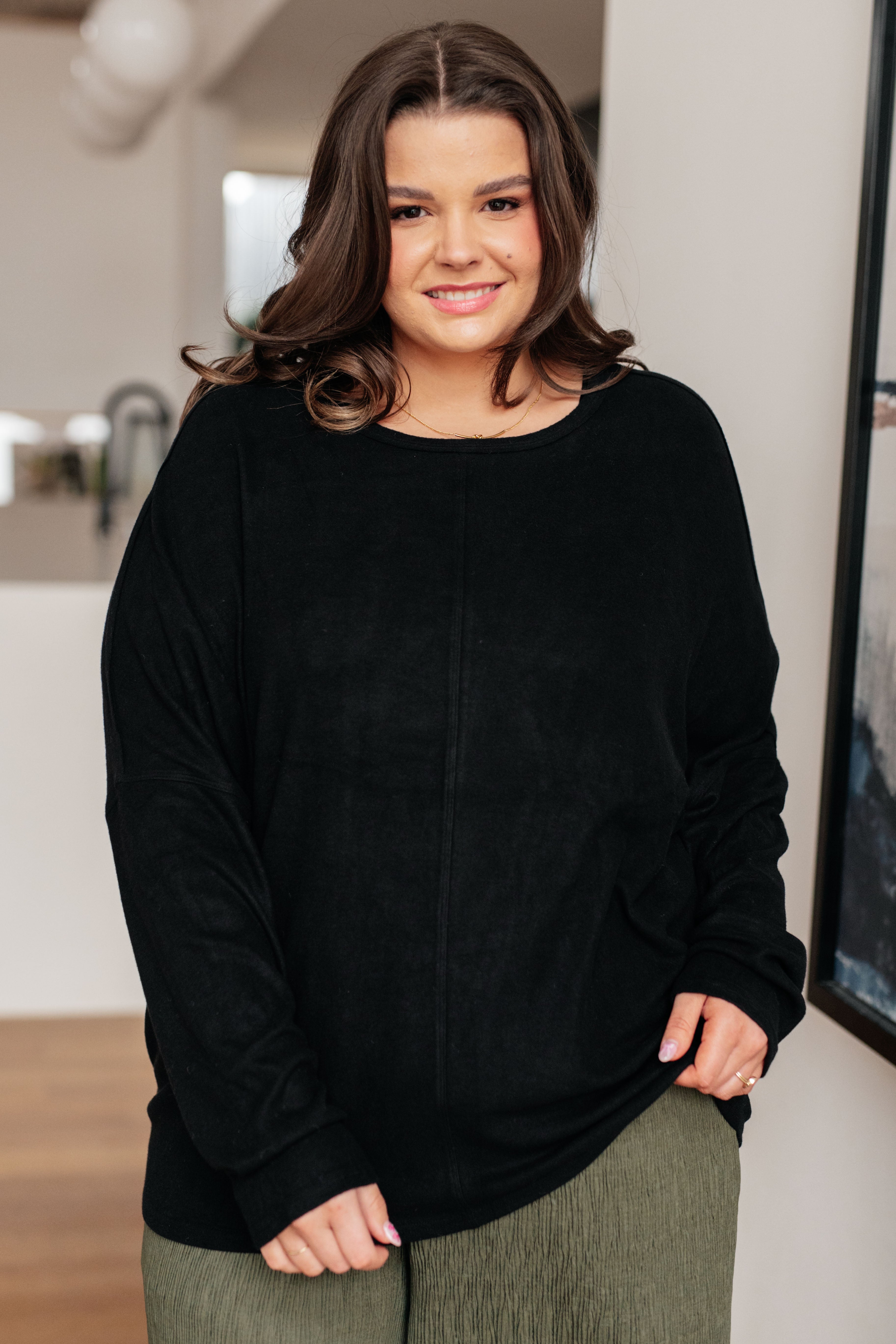 Woman wearing a black dolman long-sleeve top and green pants standing in an indoor setting.