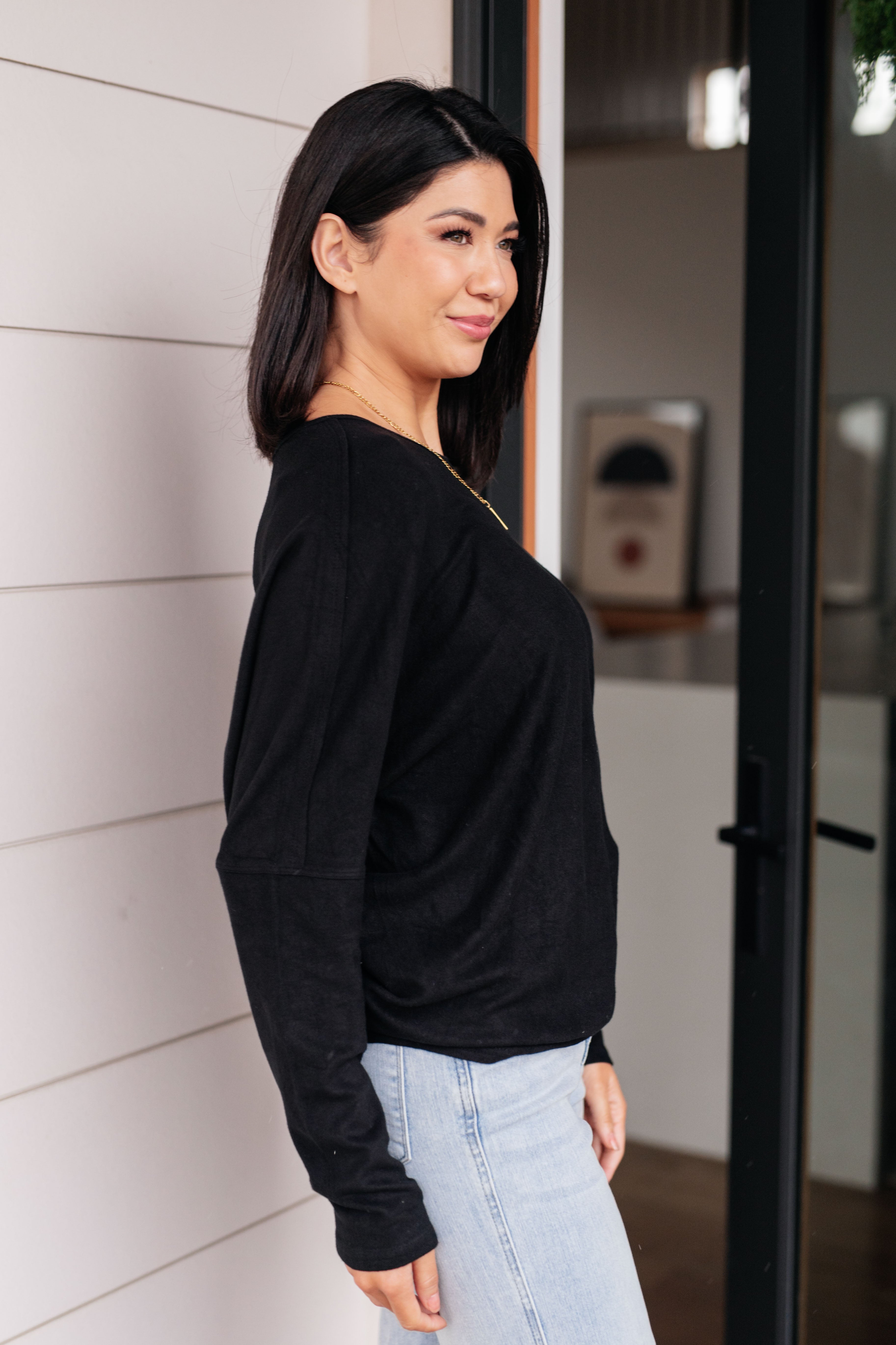 Woman wearing a black dolman long-sleeve top and light blue jeans standing in front of a glass door.